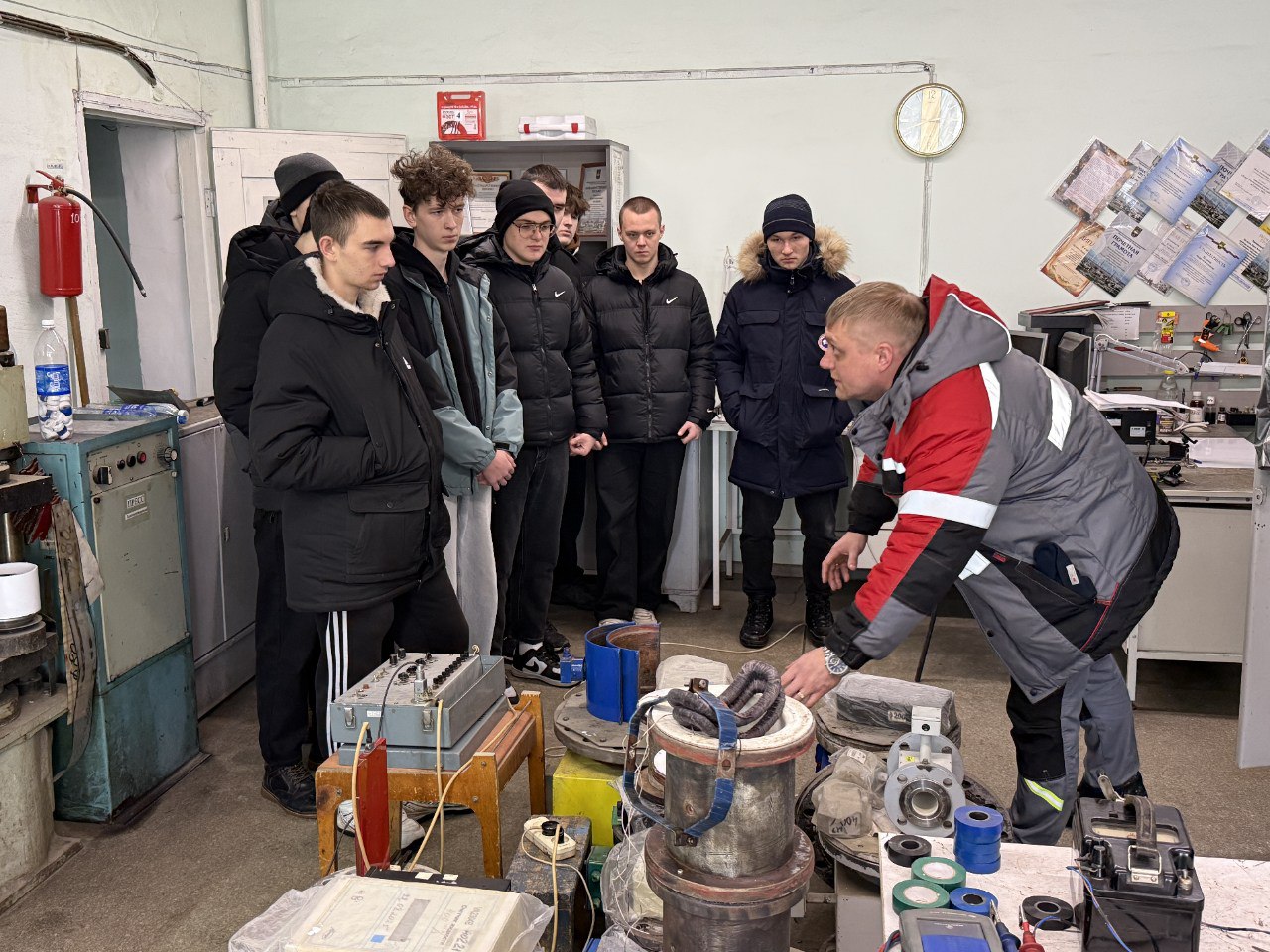 фото показывает группу из семи человек, стоящих вдоль стола, на котором размещено множество инструментов и технического оборудования. В центре стоит инструктор, объясняющий что-то группе, с жестами показывающий на приборы. Освещение в комнате хорошее, поскольку потолок светлый, а на стенах размещены различные технические документы.