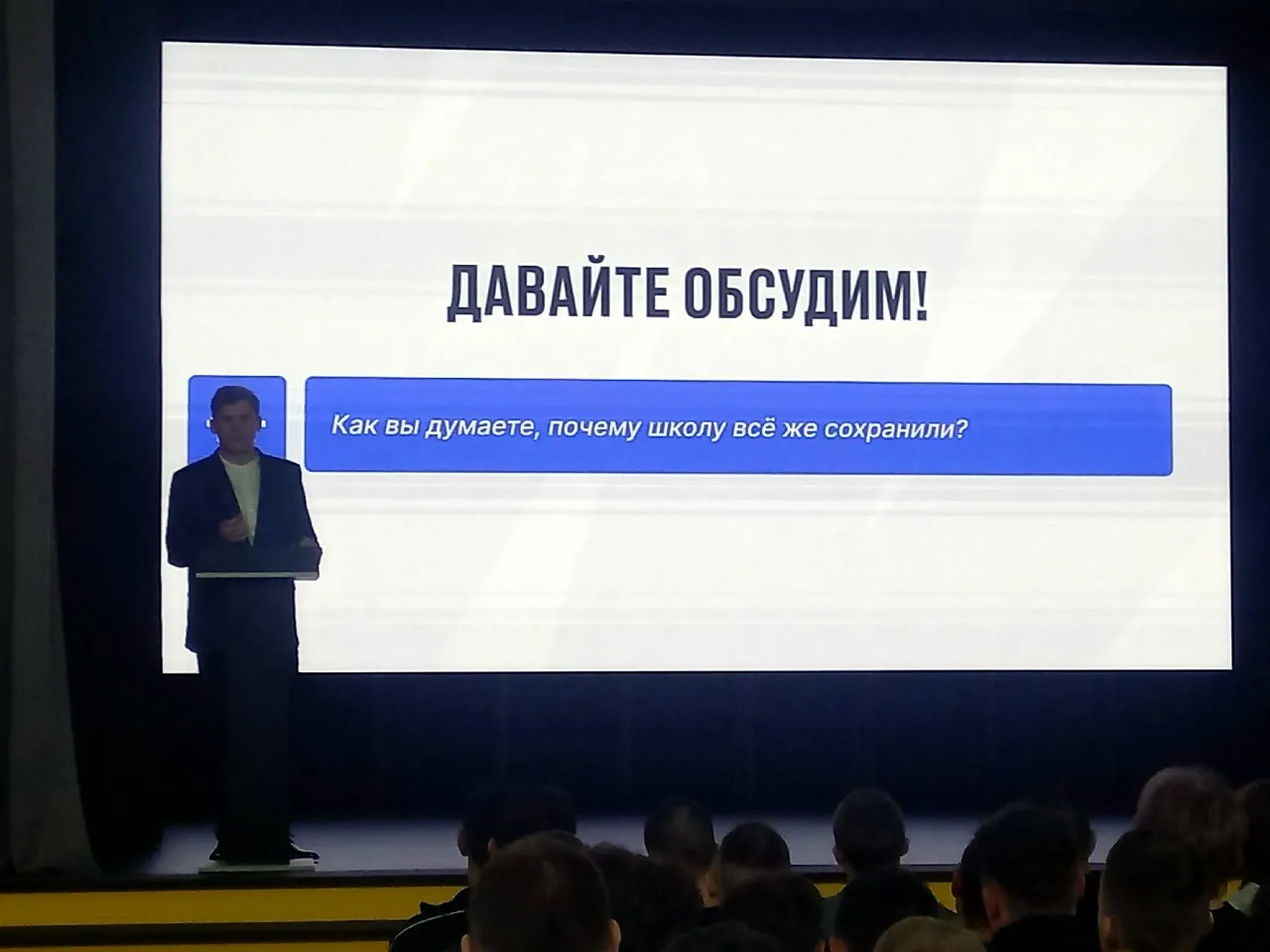 В затемнённом актовом зале на большом экране показан слайд с надписью «ДАВАЙТЕ ОБСУДИМ!» и вопросом: «Как вы думаете, почему школу всё же сохранили?». Слева у экрана стоит выступающий.