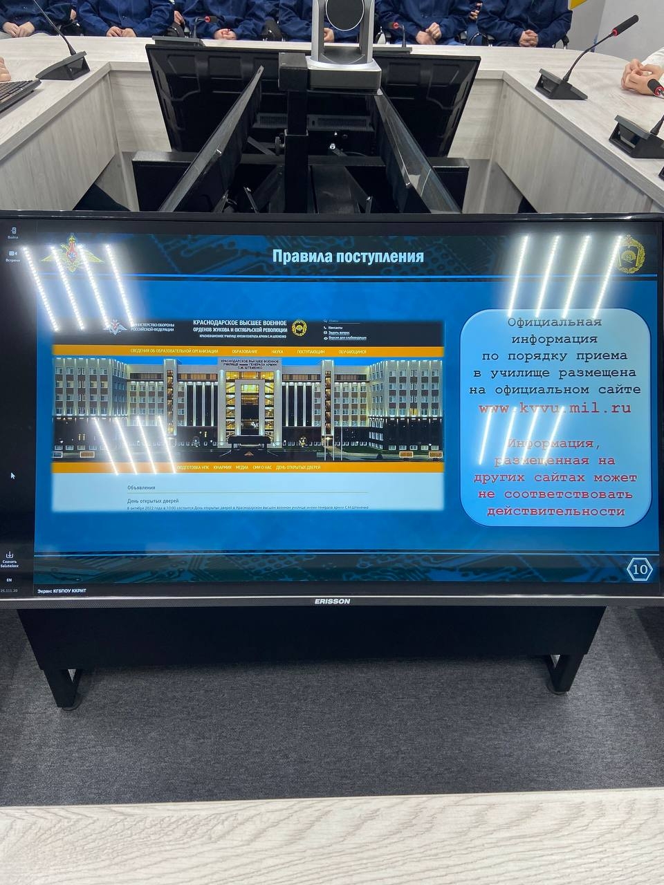 крупным планом представлена интерактивная панель или экран, установленный на столе. На экране отображается информация с заголовком «Правила поступления». Видны изображения зданий в архитектурной графике, вероятно, учебного заведения, и текст с рекомендациями по перезванию. Также видно много рабочих мест с компьютерами и микрофонами, которые расположены вокруг стола. Обстановка снова выглядит деловой, а освещение направлено на экран, подчеркивая важность представленного материала.