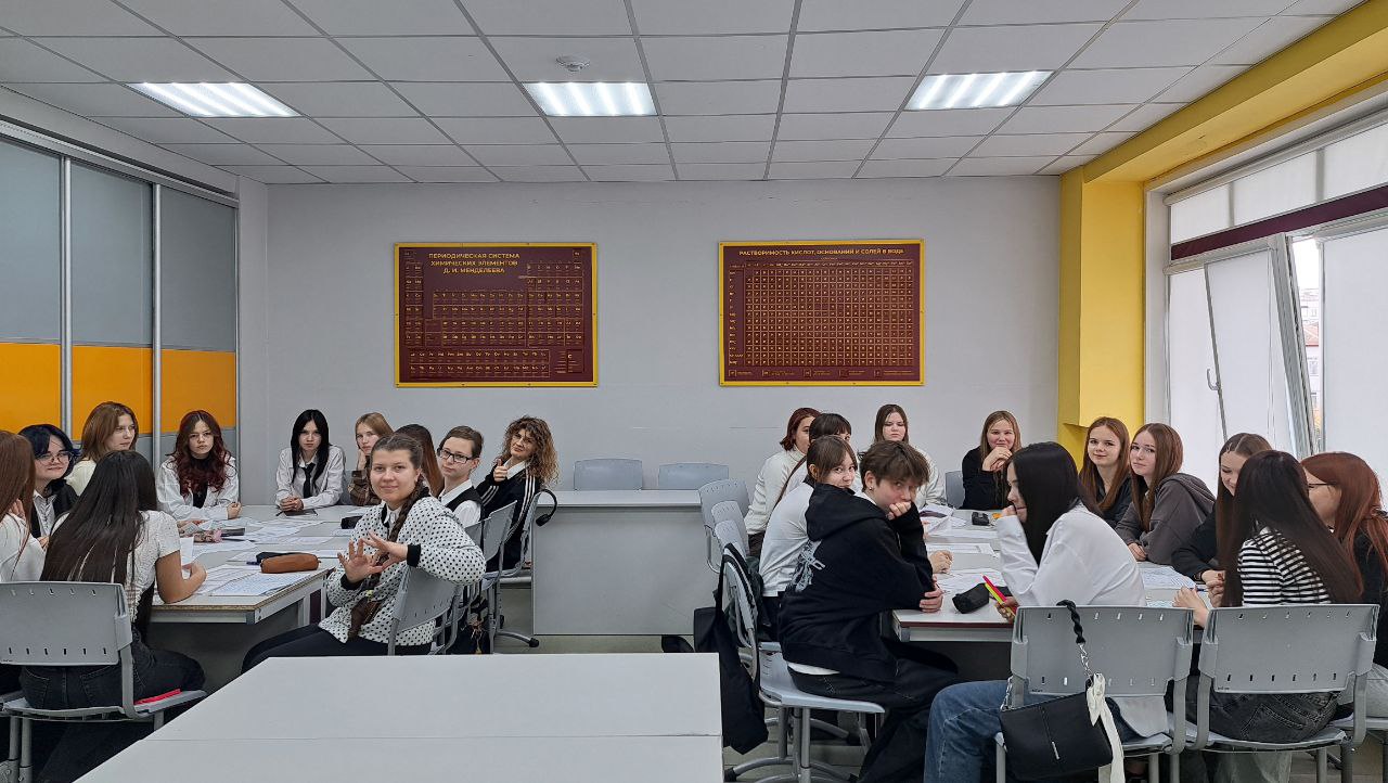 изображена группа студентов, расположившихся за длинным столом в светлом помещении. В классе видны светлые стены и желтые акценты. На стенах размещены два стенда с таблицами, возможно, с учебной информацией. Студенты сидят и общаются друг с другом, в основном, девушки, одетые в белые и черные футболки. Некоторые из них улыбаются, другие активно обсуждают что-то на столе.