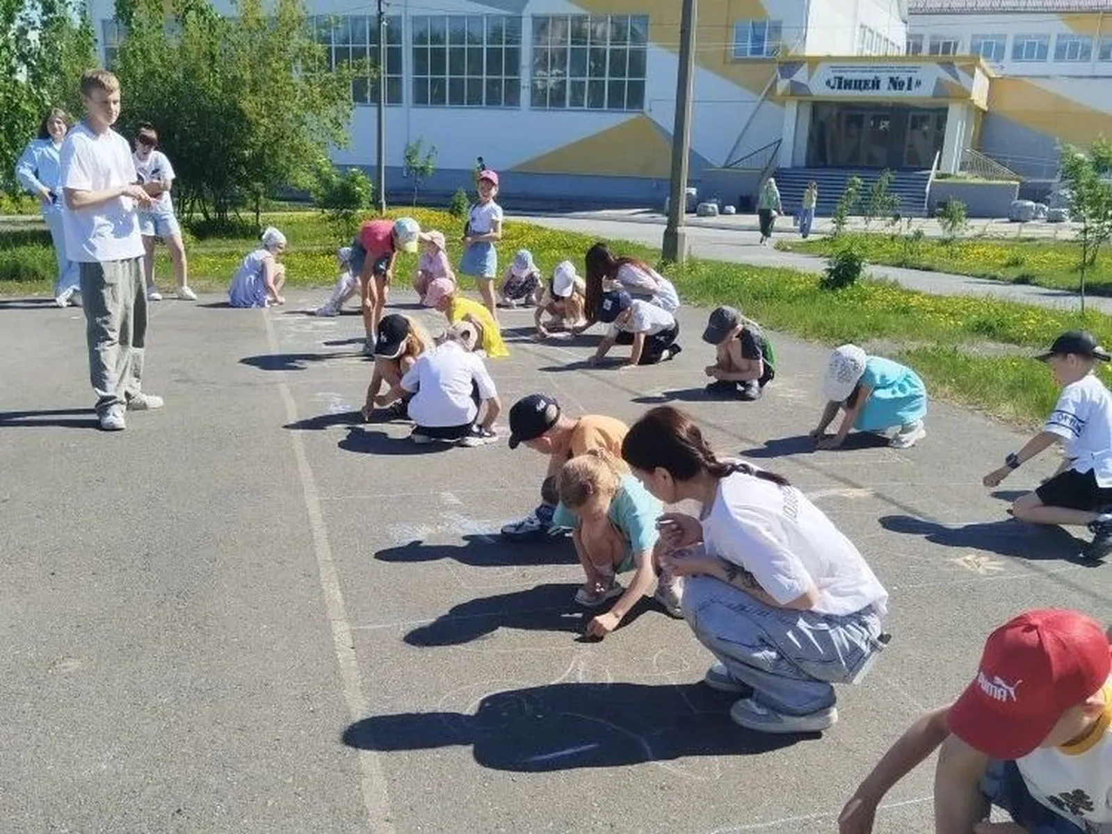 Дети рисуют мелом на асфальте: около двадцати младших детей расселись по площадке, взрослые наблюдают и помогают; здание школы виднеется позади.