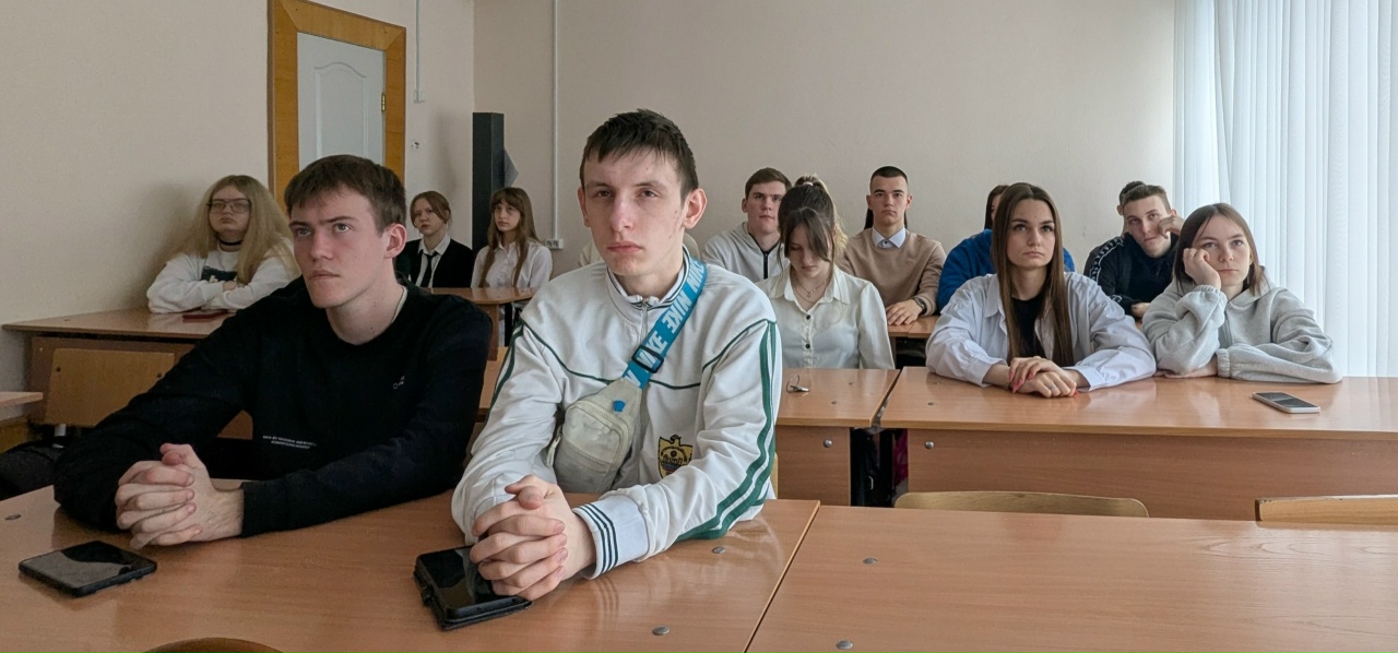 На этой фотографии показана группа студентов, которые внимательно слушают лекцию. Они сидят за столами, расположенными в ряд. Перед ними видно несколько учеников с серьезными выражениями лиц. У некоторых из них на столах лежат мобильные телефоны. Студенты одеты в светлую и тёмную одежду, что создает контраст. Позади группы видно часть классной комнаты с дверью и стойкой для оборудования, а также окно, за которым просвечивает дневной свет.