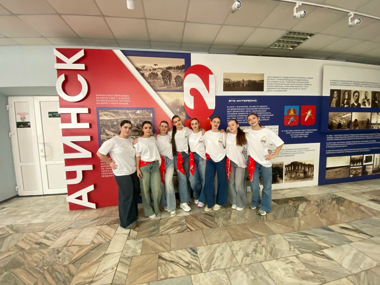 группа из восьми девушек стоит в помещении с историческим оформлением стен. Все они одеты в белые футболки и джинсы, а на талии у них завязаны красные платки. Девушки улыбаются и позируют вместе, многие слегка наклонились. За ними расположена большая панель с изображениями и текстами о городе Ачинске, включая фотографии и исторические материалы.