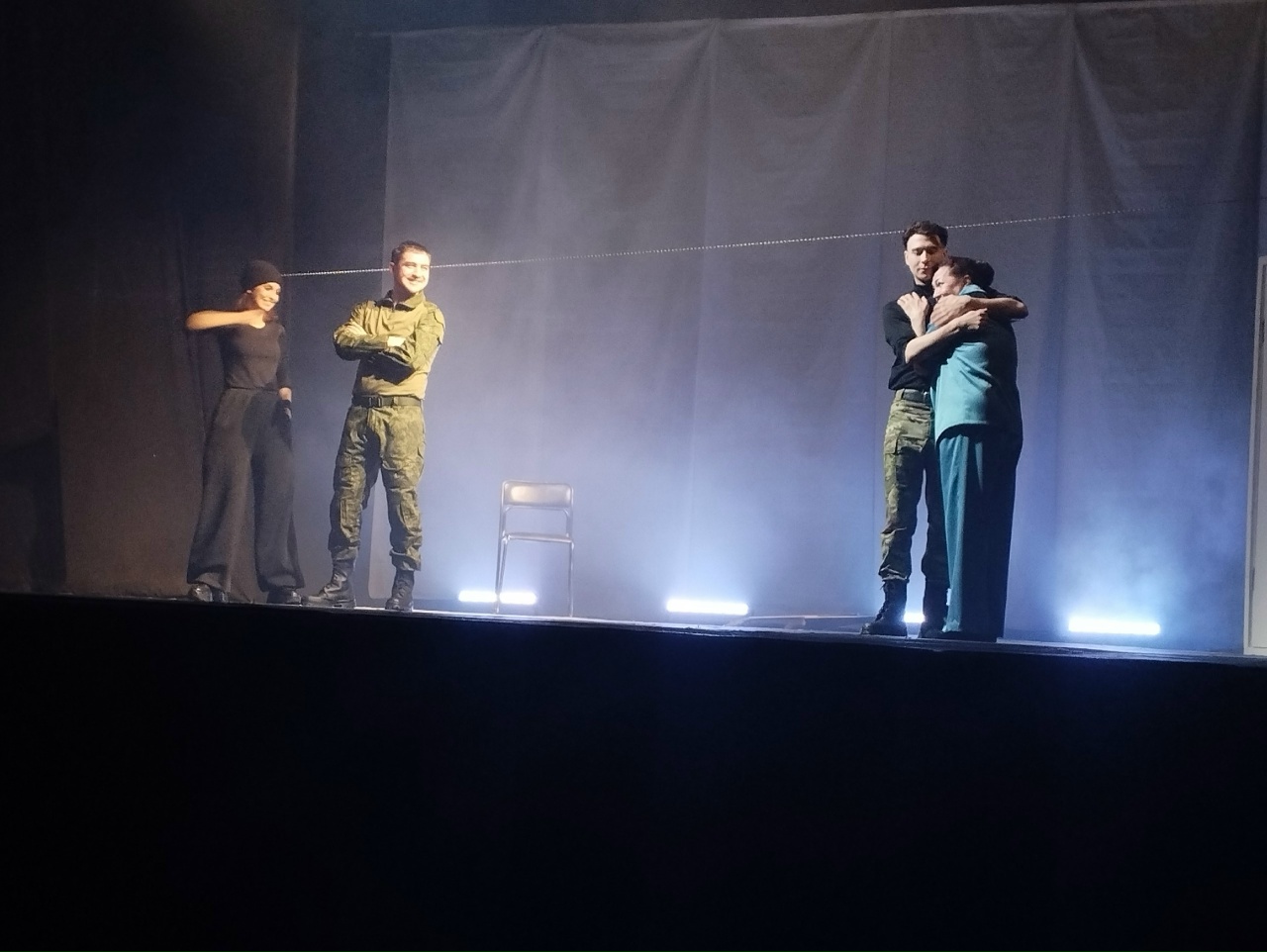 На сцене театра освещенные прожекторами стоят четыре человека, два из которых в военной форме. В центре сцены расположено пустое кресло. Один мужчина в военной форме обнимает женщину в черной одежде, а другой мужчина, также в военной форме, наблюдает за этим процессом с поднятыми руками. Атмосфера на сцене несколько эмоциональная, с легким туманом на фоне, что создает атмосферу ожидания или напряжения.