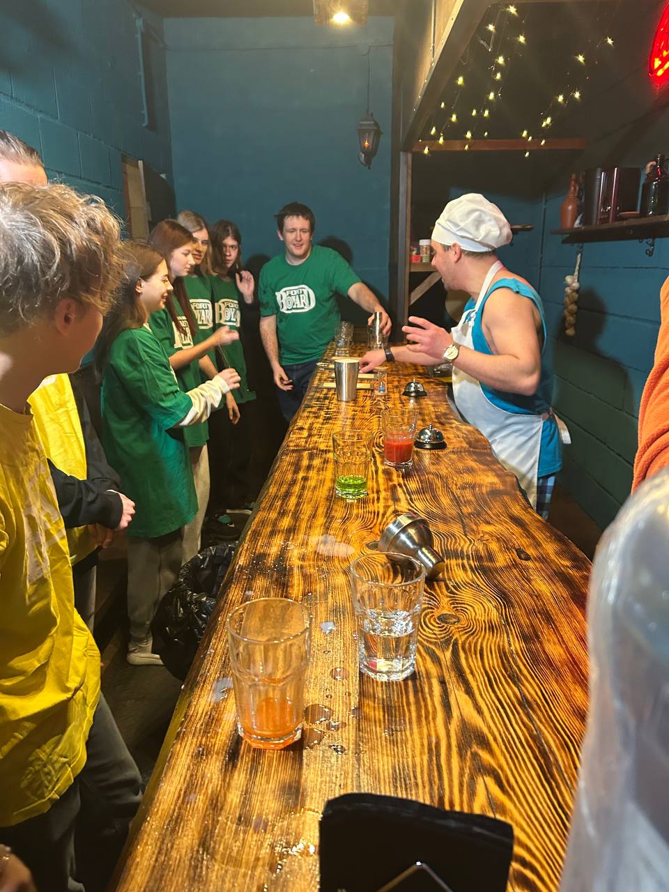 На этой фотографии изображена группа молодых людей, наблюдающих за процессом приготовления напитков. В центре стоит человек в поварском колпаке, который готовит коктейли за длинной барной стойкой. На столе присутствуют различные разноцветные жидкости, а вокруг сосредоточенные лица зрителей, которые одеты в футболки разных оттенков. В помещении мягкое освещение, создающее уютную и живую атмосферу.