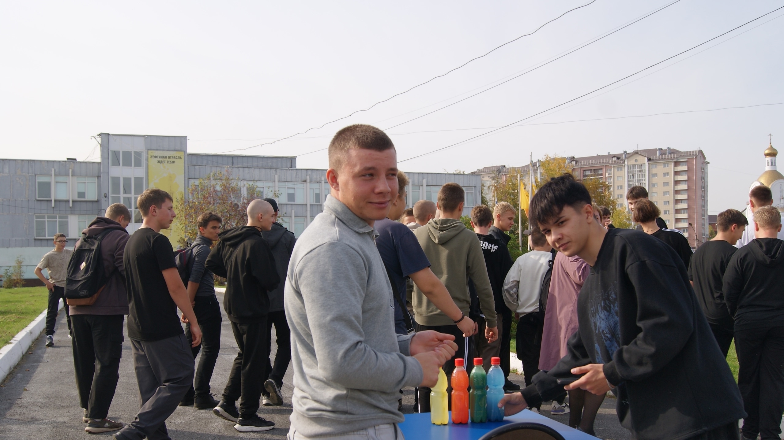 На этом снимке два молодых человека стоят за столом, на котором расположены бутылки с напитками ярких цветов. Один из них, с короткой стрижкой и в сером свитере, улыбается в камеру. Второй парень, с длинными волосами, также смотрит в объектив. На заднем плане видна группа людей, одетых в тёмную одежду.