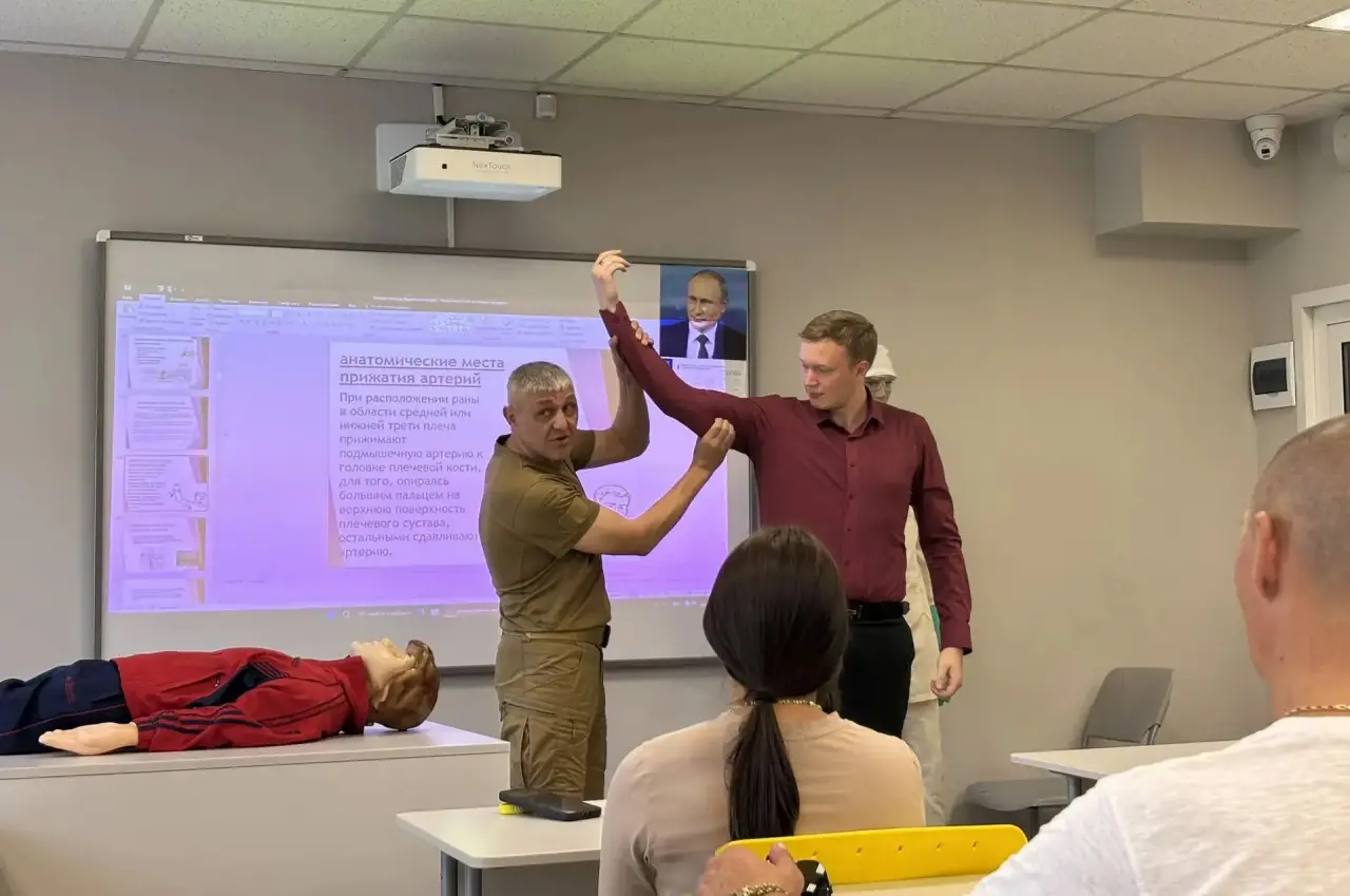 В учебном классе проходит практическое занятие по оказанию помощи. Два мужчины стоят у доски и демонстрируют прием: один держит руку другого поднятой вверх, как при обучении действиям в экстренной ситуации. Слева на столе лежит манекен или учебный пострадавший в красно-синей одежде. На заднем плане виден проектор с презентацией на экране, а в комнате сидят слушатели, наблюдающие за демонстрацией.