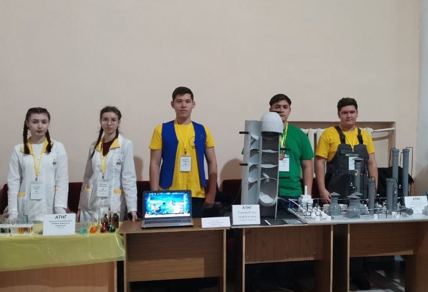 Трое молодых людей и две девушки находятся перед столом с выставочными материалами. На столе расположены разные объекты и компьютер с изображением. Один из юношей присутствует в желтой футболке и синем жилете, а остальные – в белых и серых униформах. Все они стоят на фоне стенда, на котором видно название их команды или проекта. Обстановка выглядит оживленной, возможно, идет подготовка к защите проекта или демонстрации.