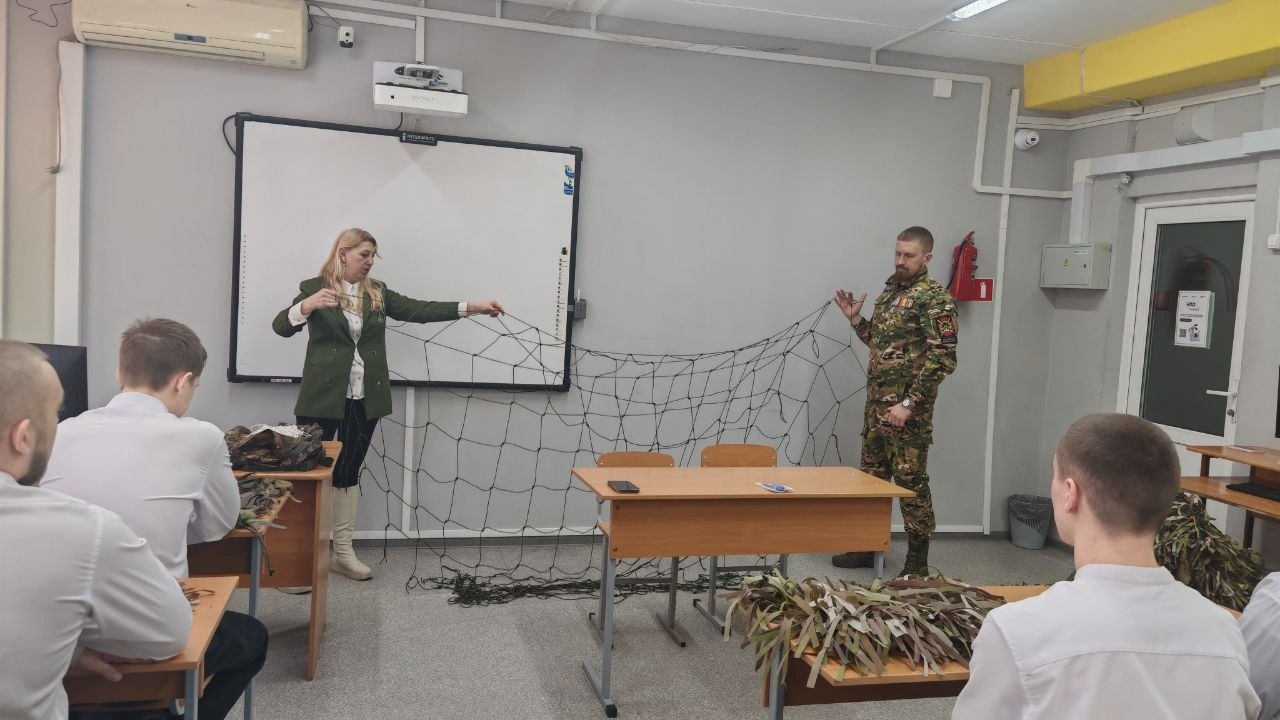 запечатлен класс, в котором два человека демонстрируют сетку. Женщина стоит слева, одетая в светлый блузон и пиджак. Она держит сетку в руках, показывая ее структуру. Справа от нее стоит мужчина в камуфляжной форме, который также держит часть сетки. На столе перед ними расположены несколько неровно сложенных веток и листьев, что создает ощущение природного окружения. В аудитории видно группу студентов в белых рубашках, которые внимательно смотрят на демонстрацию.