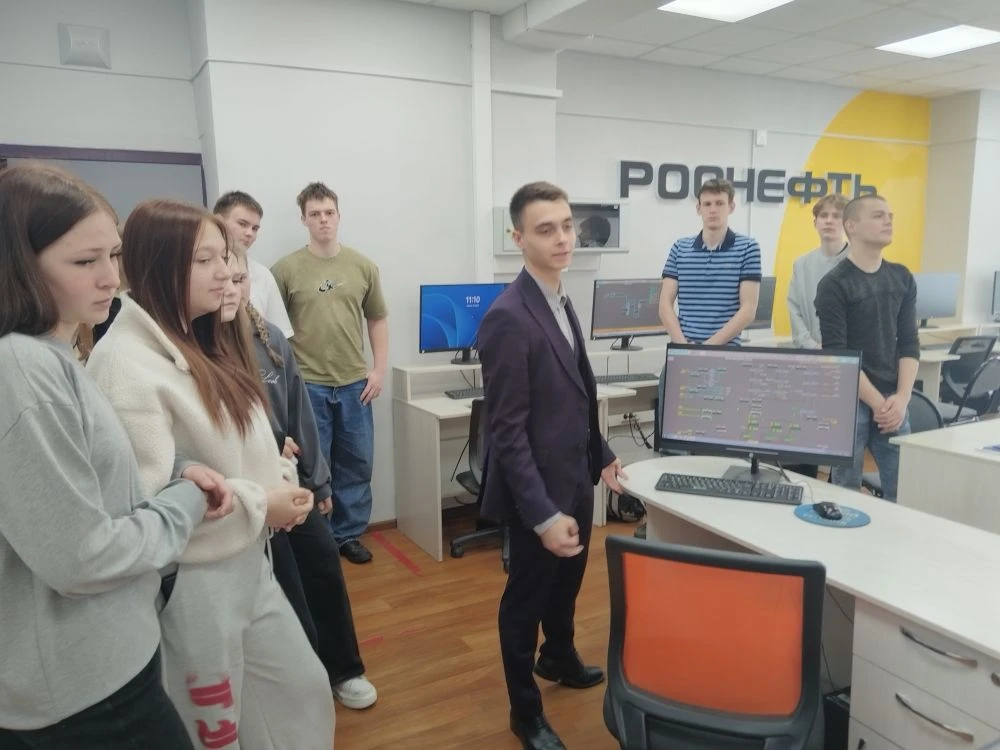 изображена группа людей, стоящих в комнате с компьютерами. Спереди стоит молодой человек в темном костюме, который, похоже, демонстрирует экран компьютера. Позади него находится группа из шести человек, включая мужчин и женщин, одетых в повседневную одежду. Обстановка выглядит современной, с яркими цветами и технологичным оборудованием.