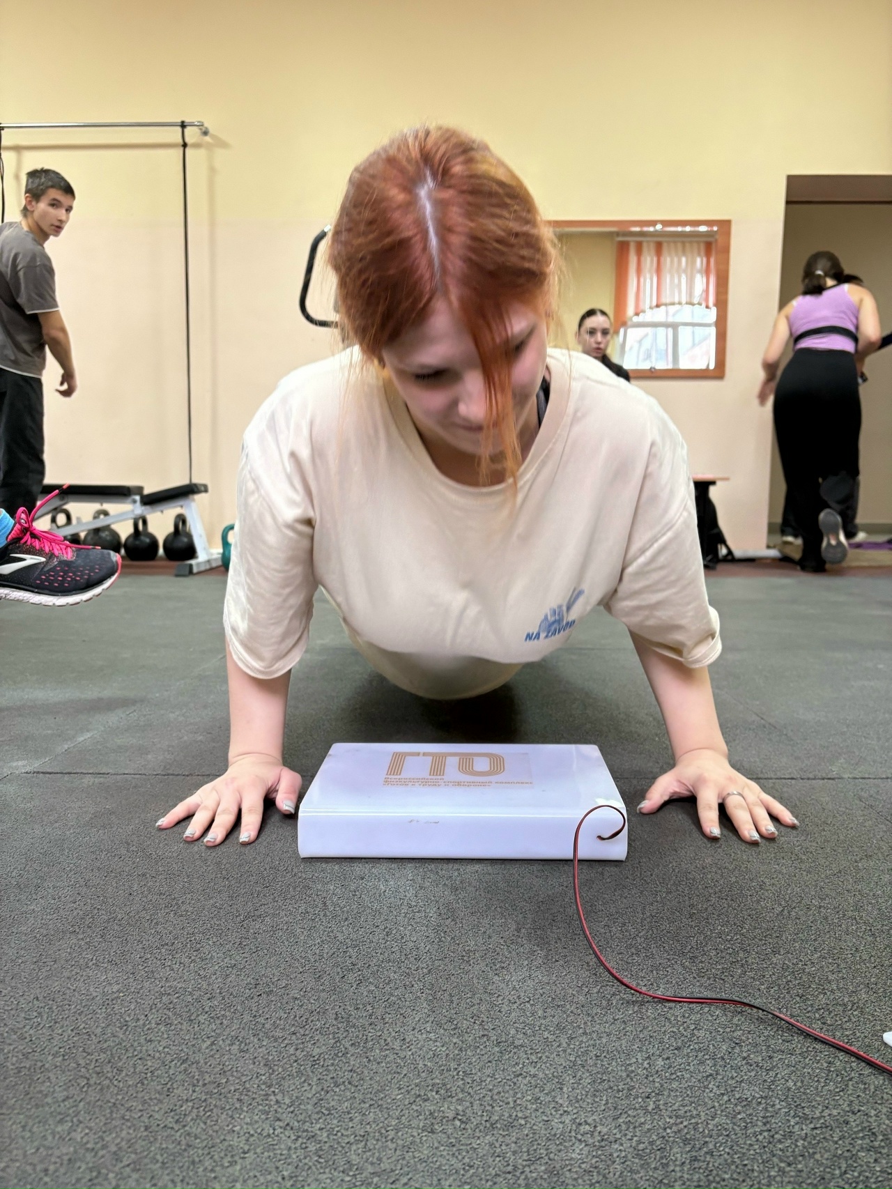 На изображении фигура девушки с длинными рыжими волосами, которая находится в положении упора на руках, выполняя физическое упражнение — планку. Она одета в светлую футболку и сосредоточена на выполнении упражнения. На полу видно спортивный инвентарь, а в фоновом пространстве находятся другие занятия, где тренируются люди.