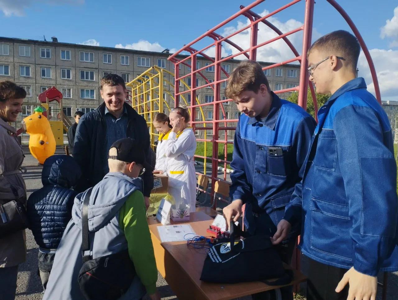 Люди за столом на уличной площадке во время мастер-класса или выставки: двое мужчин в синих рабочих куртках стоят у стола с инструментами и проводами, рядом дети и взрослые, на заднем плане — игровые и спортивные конструкции.