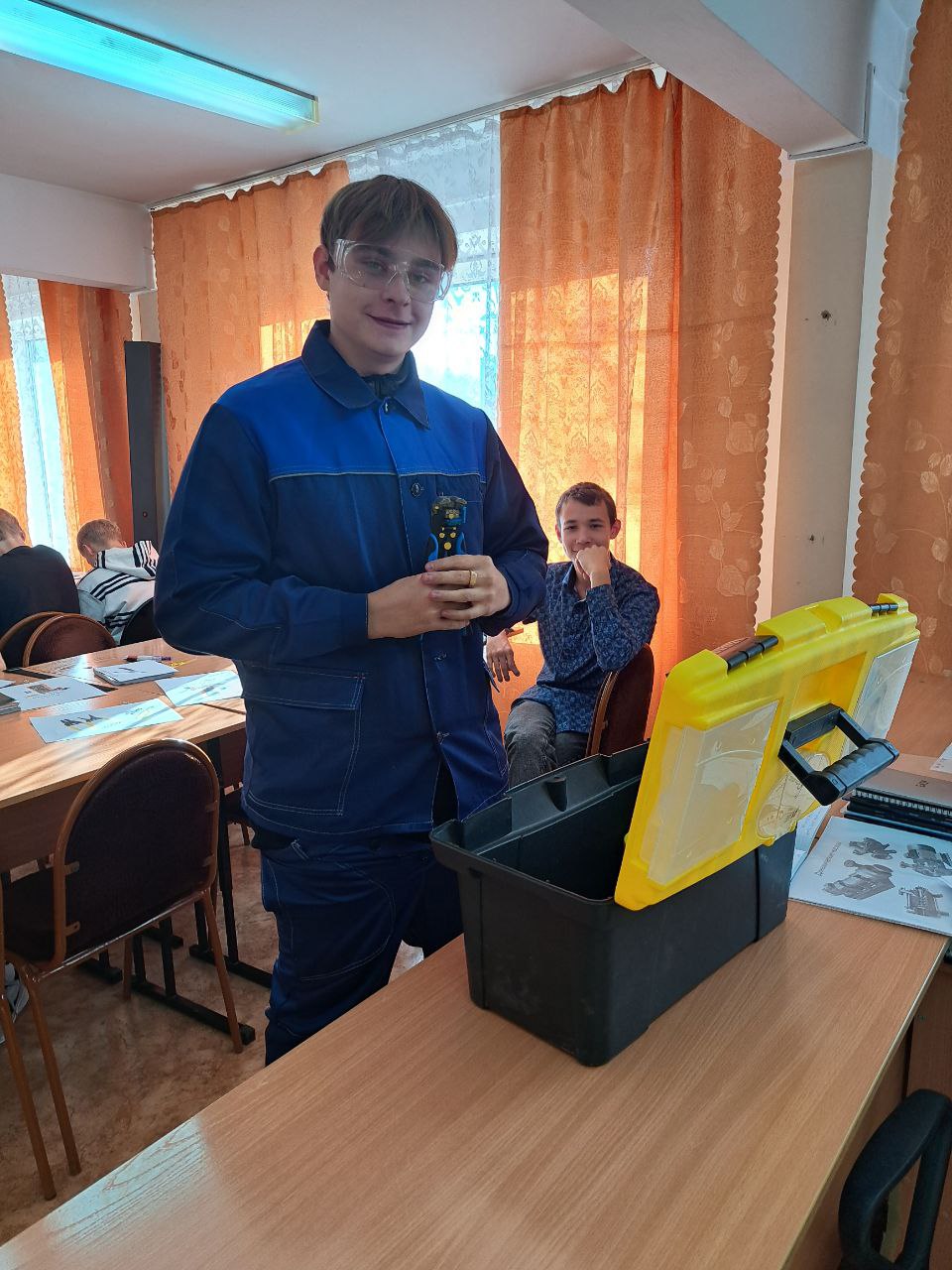 На переднем плане стоит юноша в синем рабочем комбинезоне и очках. Он улыбается и держит в руках какой-то предмет, возможно, инструмент. За ним видно несколько людей, сидящих за столами, на которых лежат листы бумаги. Оконные шторы оранжевого цвета пропускают немного света.