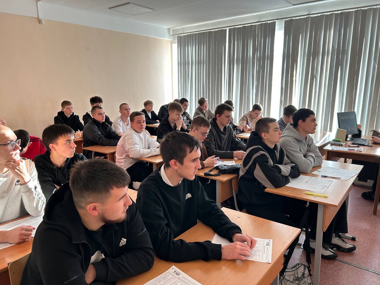 На этом фото изображена другая классная комната, в которой ученики слушают занятие. Существует около двадцати учеников, большинство из которых одеты в темную спортивную одежду. Они сидят за партами, некоторые из них склонили головы, обдумывая информацию, а многие смотрят вперед на учителя или экран.