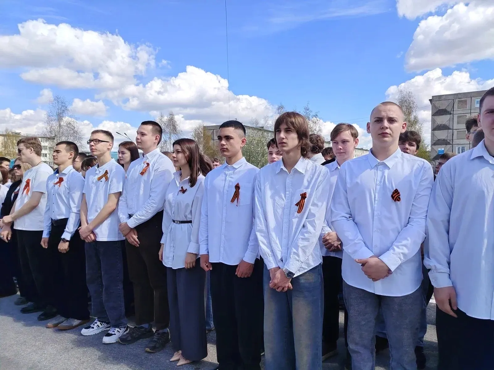 На фото изображение группы молодых людей, стоящих в ряд на улице. Все они одеты в белые рубашки, некоторые из них имеют на себе георгиевские ленты. Впереди стоит мужчина с короткой стрижкой, а также несколько девушек и юношей. На заднем плане видны облака и голубое небо, а также здания, что указывает на то, что мероприятие проходит на открытом воздухе.