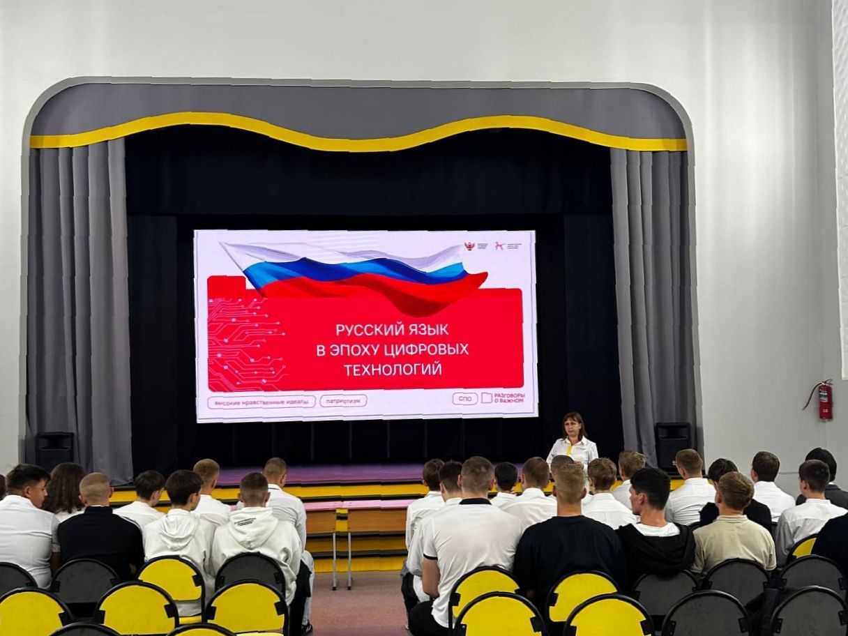 На сцене находится большой экран, на котором отображается текст "РУССКИЙ ЯЗЫК В ЭПОХУ ЦИФРОВЫХ ТЕХНОЛОГИЙ". Перед экраном сидит группа студентов, одетых в белые футболки. Сцена оформлена в светлых тонах с желтыми и черными элементами. Атмосфера выглядит учебной и сосредоточенной.