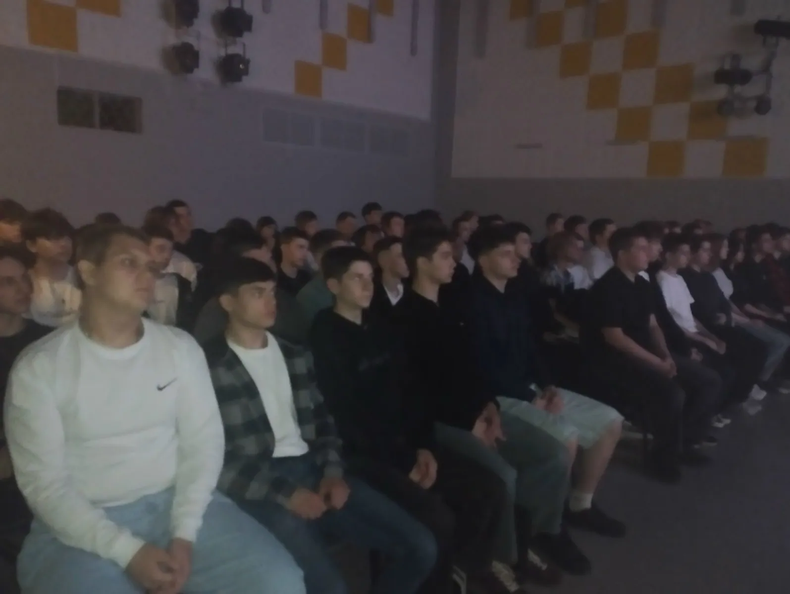 Зал с аудиторией: тёмный кинозал/актовый зал, ряды сидящих — преимущественно молодые мужчины и подростки, смотрящие вперёд на экран; видны несколько рядов в анфас, низкая освещённость.