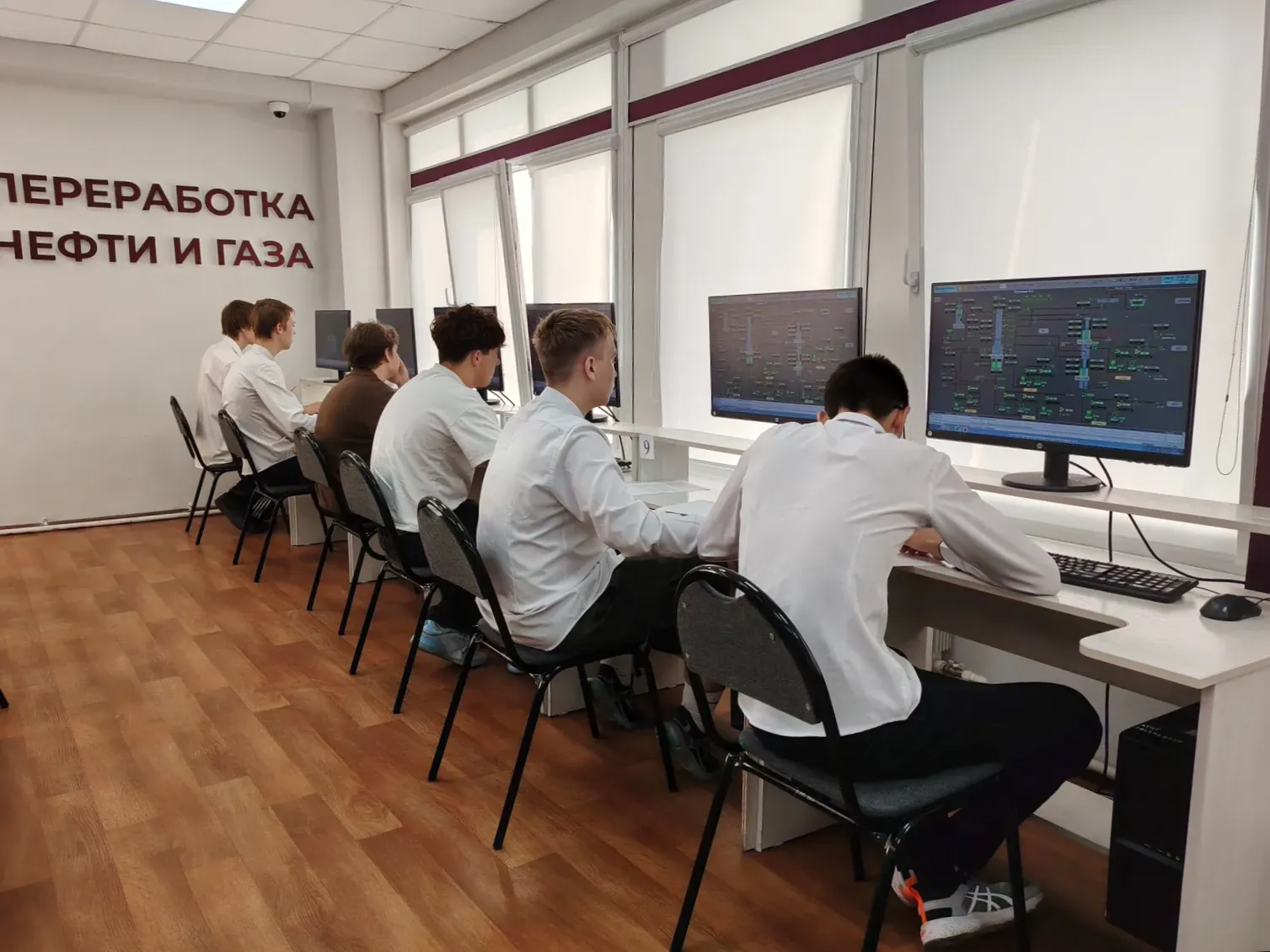 Классная комната с рядами компьютеров у стены: несколько студентов сидят за мониторами и работают за экранами с техническими схемами; надпись на стене о переработке нефти и газа.