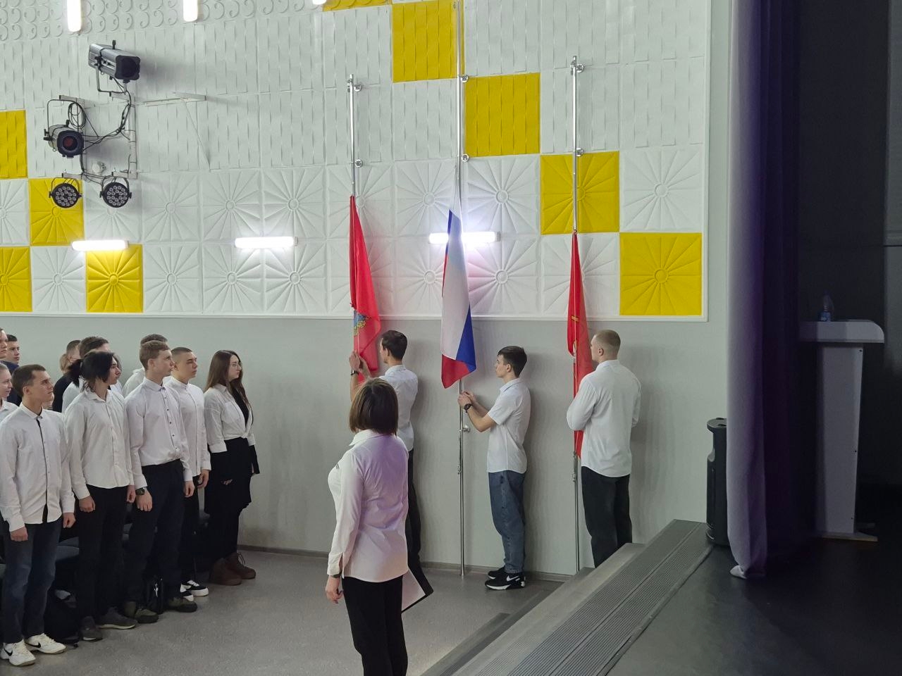 На фото группа молодых людей стоит в форме полукруга, обращенная к флагам и сцене. Два молодых человека, одетые в светлые рубашки, готовятся установить флаги. Женщина из первой фотографии стоит перед ними с документами в руках. Справа на фото видны серые стенки и пульт управления.