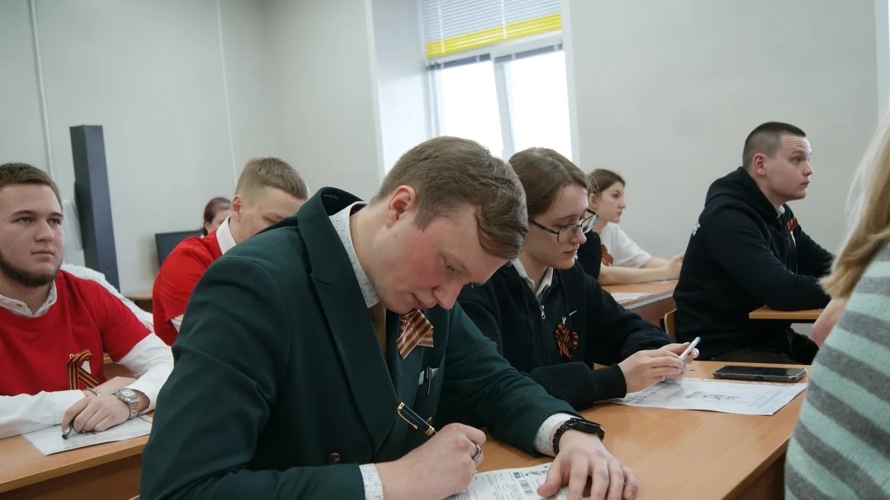 показан класс, но фокус сделан на нескольких студентах. Справа сидит молодой человек в темно-зеленом пиджаке, который внимательно записывает что-то в блокнот. Рядом с ним сидит девушка в светлом свитере, которая также делает записи. На заднем плане видно еще нескольких студентов в черной и красной одежде, некоторые из которых тоже носят георгиевские ленточки. Светлые стены и окна создают яркую атмосферу.