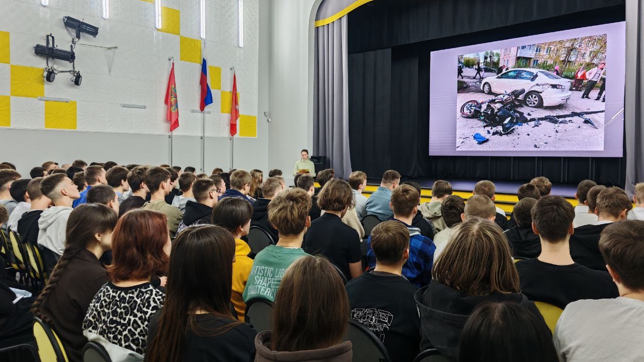 На экране показано изображение автомобильной аварии: мотоцикл и автомобиль, рядом разбросанные детали. Перед экраном находится спикер, который, вероятно, объясняет ситуацию. Аудитория состоит из молодых людей, внимательно смотрящих на экран. В зале висят государственные флаги, а также видно освещение, создающее атмосферу для лекции.