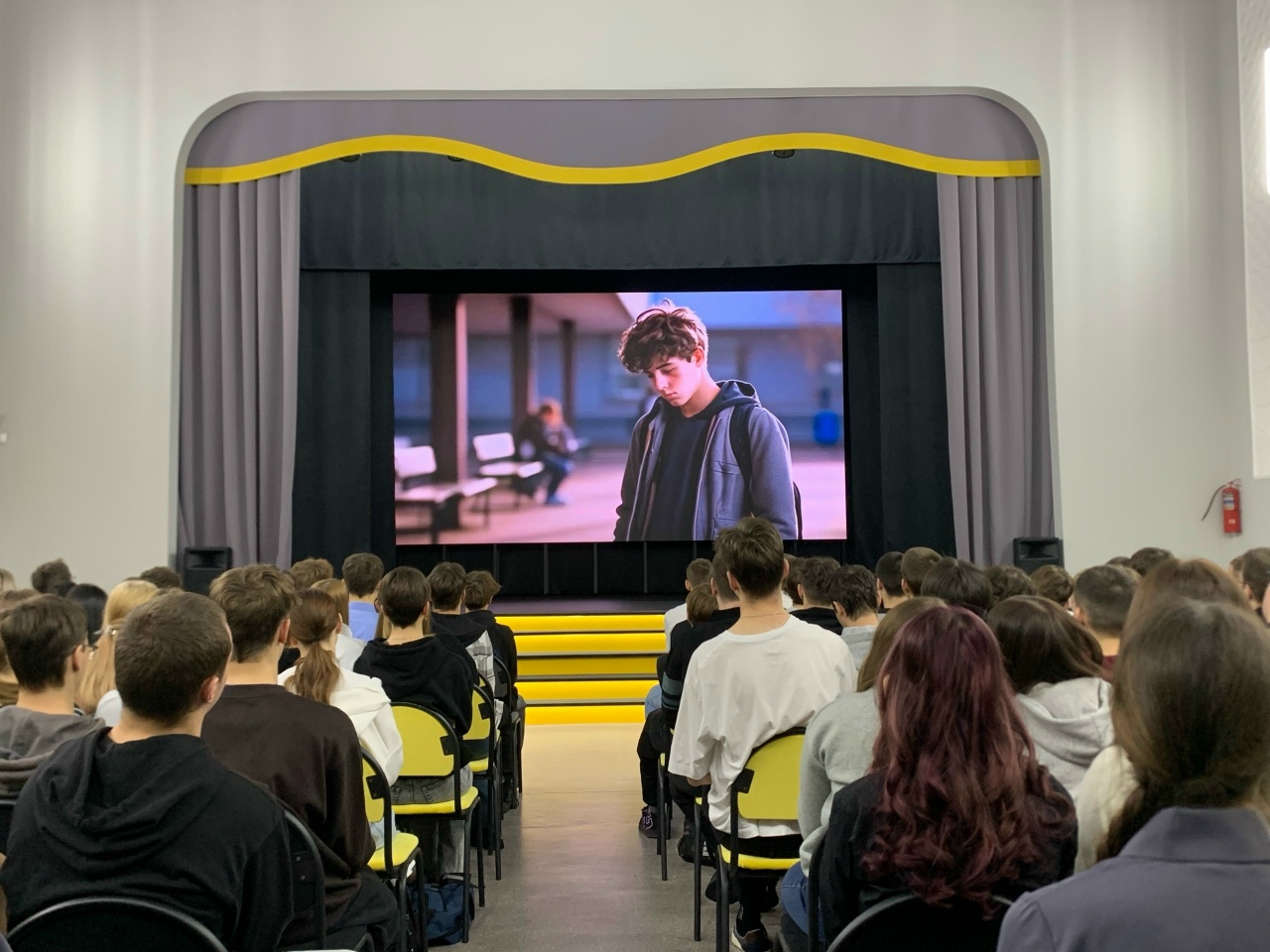 показан экран, на котором изображена сцена с молодым человеком, стоящим в помещении, возможно, в аэропорту или на вокзале. Оглядка вокруг говорит о том, что он может быть в состоянии раздумий или беспокойства. Аудитория по-прежнему видна на фоне, и они внимательно смотрят на экран, освещая атмосферу сосредоточенности на происходящем.