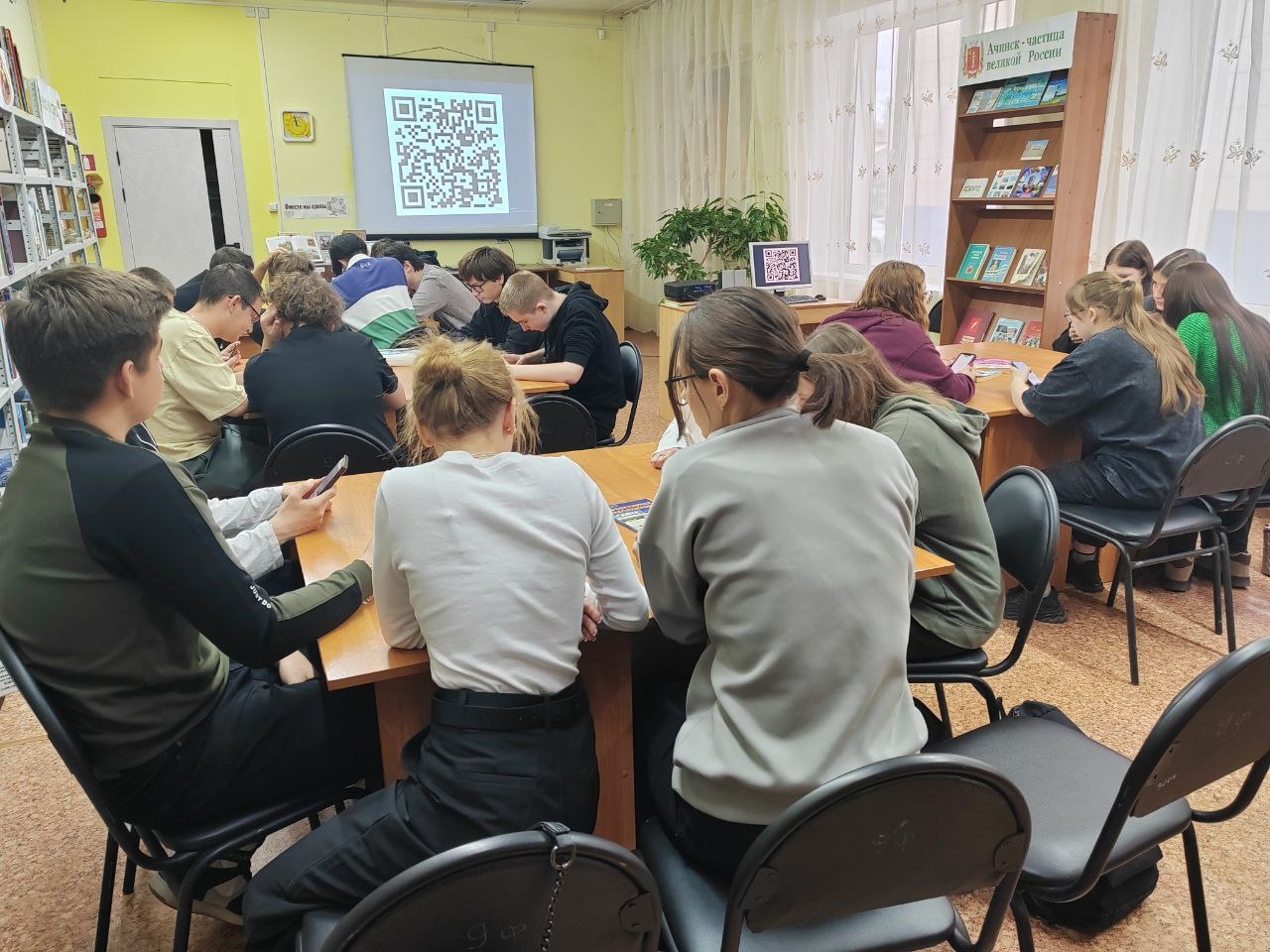 Это изображение группы студентов, которые расположились за длинным столом в библиотеке. Большинство из них смотрят в свои телефоны или папки, возможно, выполняя задания или ищя информацию. На стене проецируется QR-код, что может указывать на использование технологий в обучении. В комнате светло и уютно, что создает комфортную обстановку для учебного процесса.