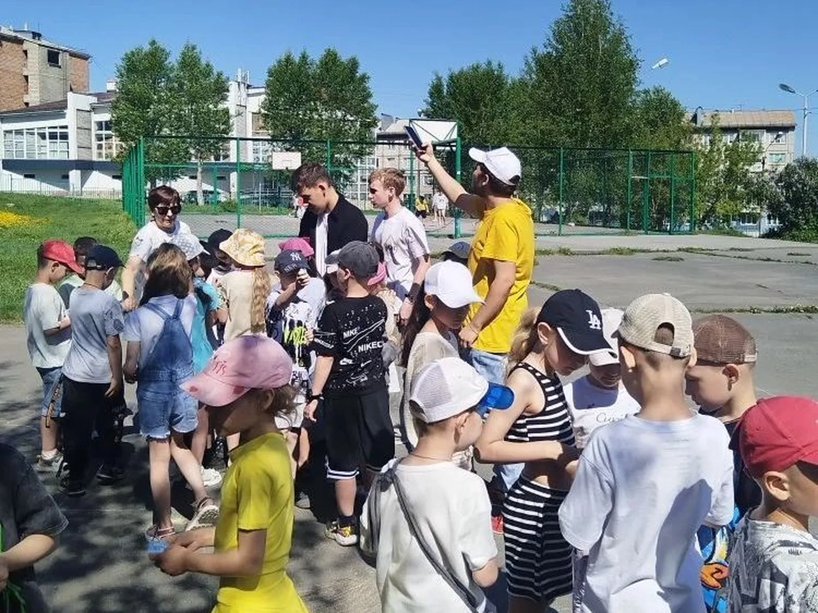 Сбор детей на улице: группа детей в летней одежде и кепках стоит вокруг нескольких взрослых (один в жёлтой футболке), взрослые дают инструкции на школьном дворике.