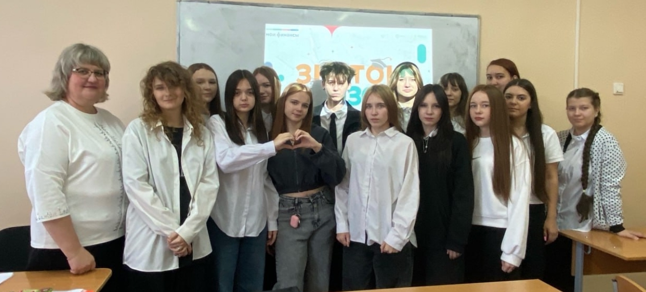 группа девушек, их 16, стоит в классной комнате. Они одеты в черно-белую одежду. В центре видно, как одна из девушек делает жест "сердце" руками. На доске также отображается слайд с надписью “Знаток”. Участницы выглядят счастливыми и расположенными, их волосы различной длины и цвета делают группу разной, но гармоничной. Атмосфера позитивная и теплоя.