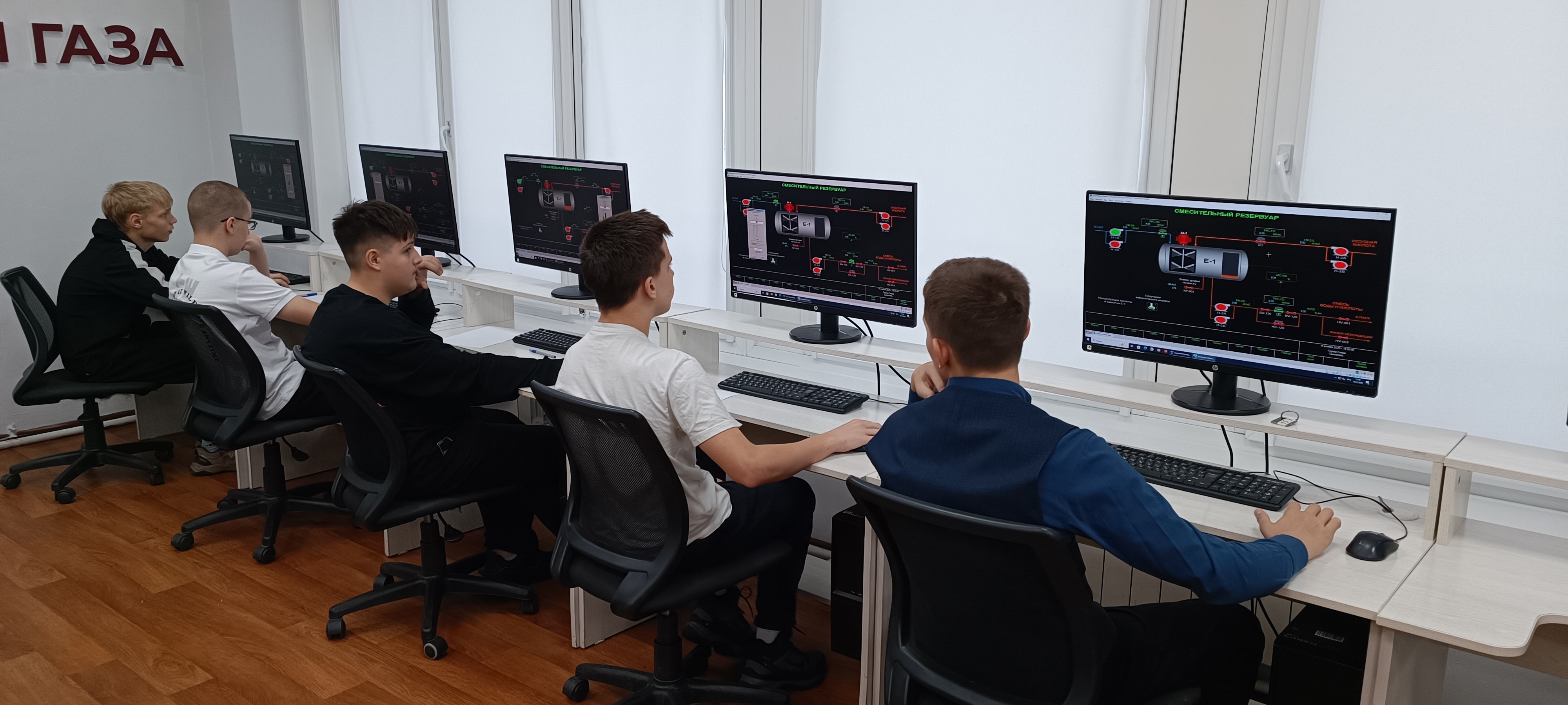 Группа студентов сидит за рядом расположенными компьютерами. Каждый из них внимательно смотрит на экраны, погруженный в процесс изучения. Студенты располагаются близко друг к другу, создавая ощущение коллективной работы. Светлая комната с деревянным полом дополнительно подчеркивает уютную образовательную обстановку.