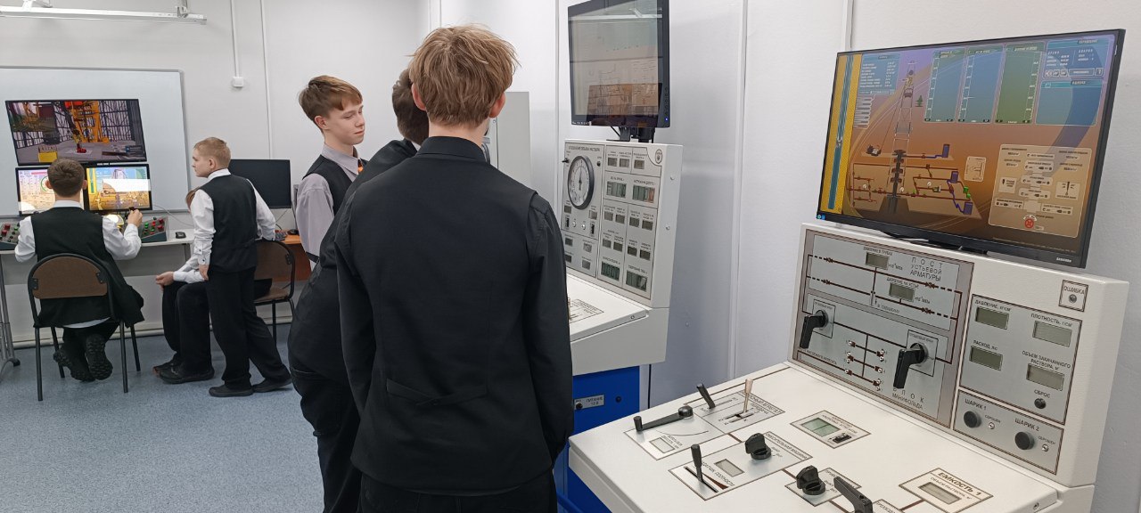 мальчики стоят у стенда с управленческой панелью. На экранах, расположенных над панелью, можно увидеть графические интерфейсы с разными контрольными функциями. Некоторые подростки смотрят на экраны, а другие - на панели управления. Окружение выглядит образовательным и технологическим, что свидетельствует о практическом обучении в технической области.
