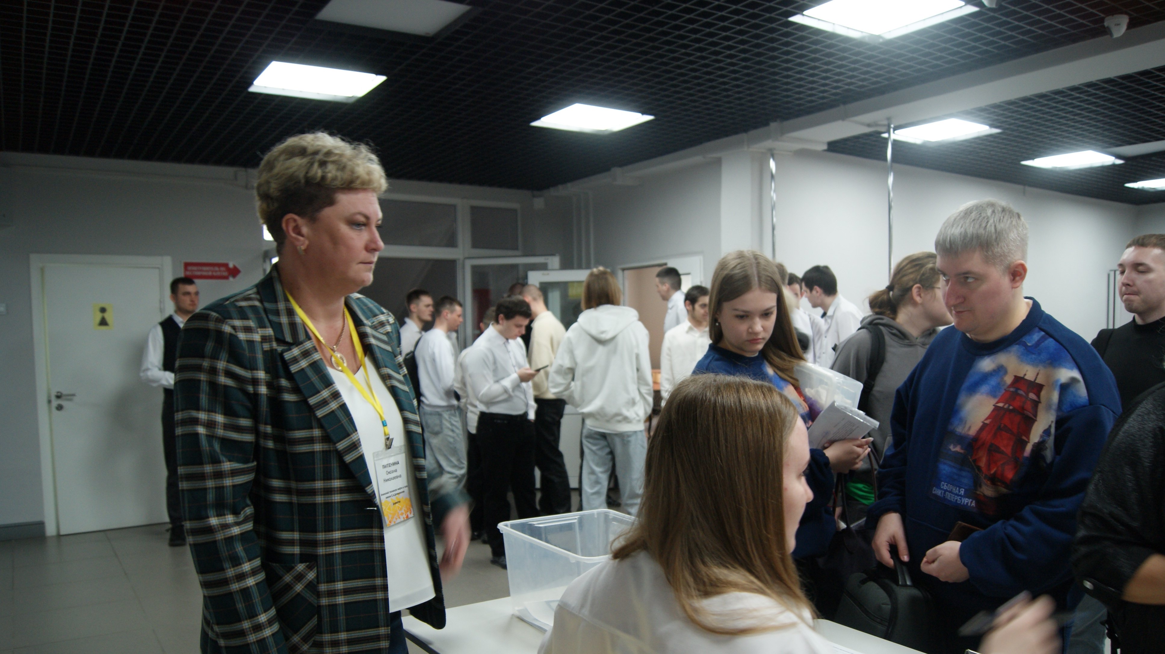 Помещение с группой людей: в центре слева — женщина в клетчатом пиджаке и ярком бейдже, вокруг неё несколько стоящих и ожидающих молодых людей; на заднем плане видна очередь и двери с указателями.