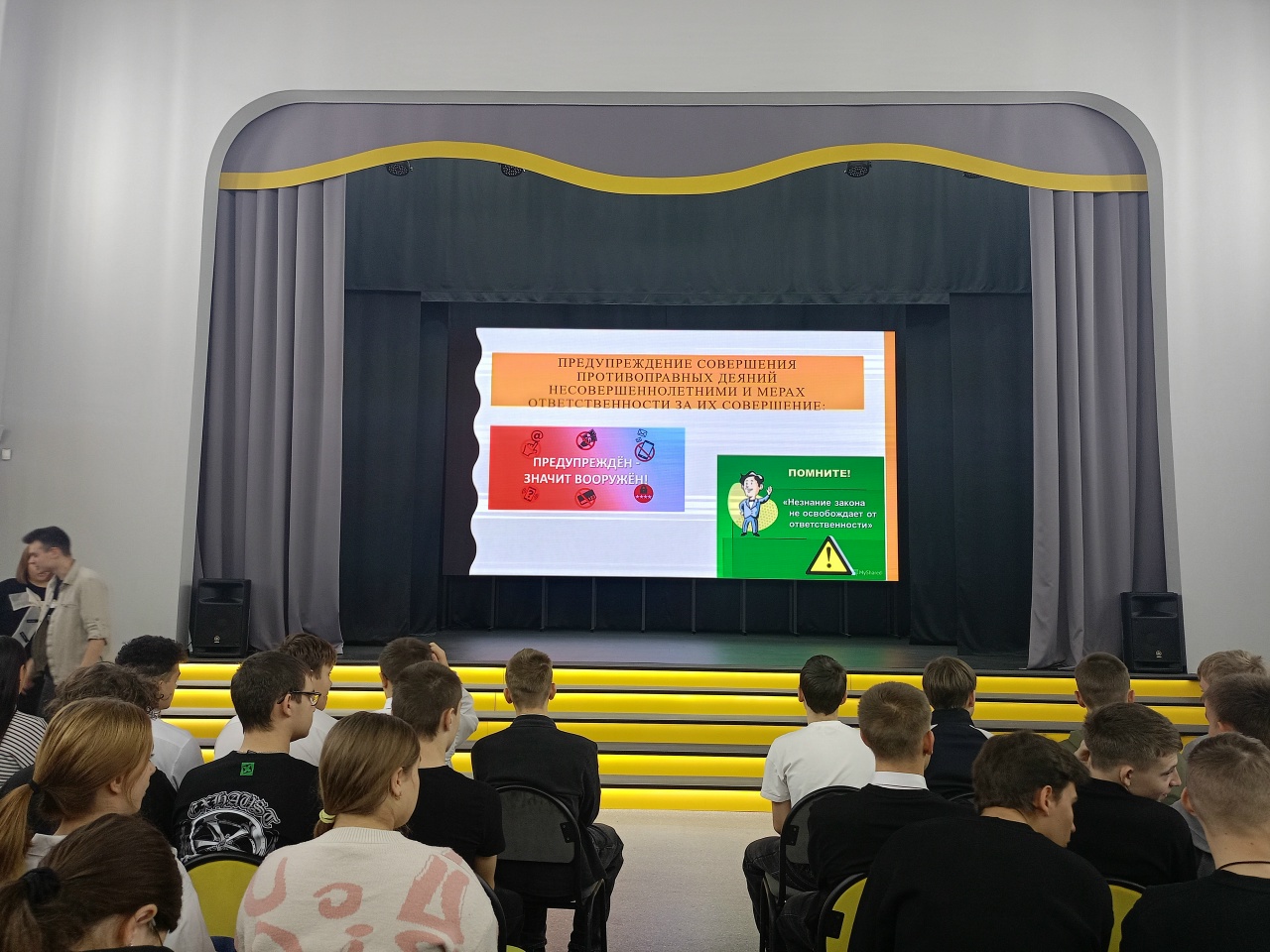 представлена ещё одна лекция, где на экране демонстрируется информационный слайд с яркими графиками и текстом. На экране видны символы, указывающие на необходимость соблюдения мер безопасности и охраны труда. В аудитории также много студентов, внимательно слушающих. Лектор, скорее всего, делится важной информацией, что подчеркивают цветные графические элементы, привлекающие внимание участников.