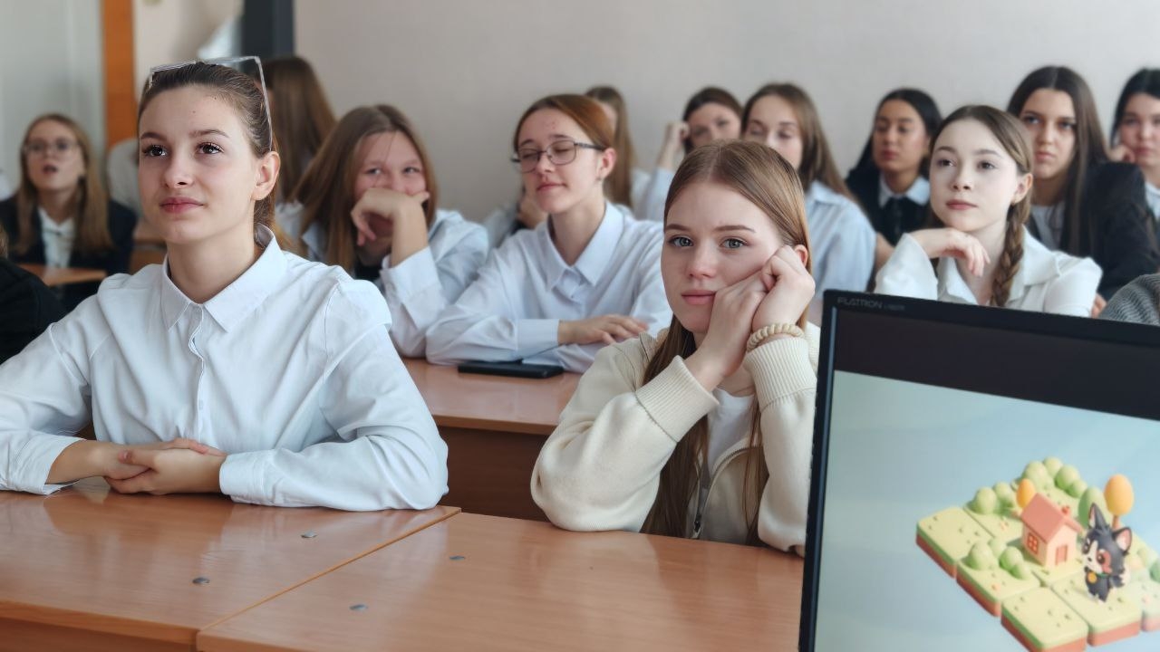 На первом фото изображены ученицы в классной комнате. Взгляд всех девушек направлен вперед. Они сидят за длинным столом, одеты в белые рубашки. Некоторые из них имеют длинные волосы. Атмосфера кажется сосредоточенной, как будто они внимательно слушают урок или презентацию.