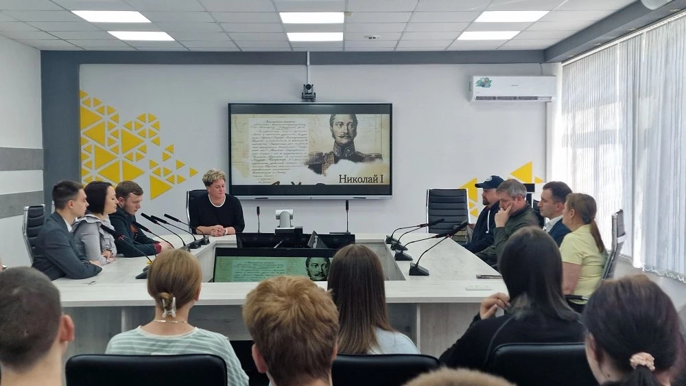 фото группа людей сидит за длинным столом в уютном помещении. В центре расположена большая телевизионная панель, на которой отображается изображение исторической личности — вероятно, Николаю I, поскольку видно текст, связанный с ним. Одна из женщин в центре стола, вероятно, ведет обсуждение или лекцию. Присутствующие активно слушают и взаимодействуют друг с другом, создавая атмосферу обучающего мероприятия. Помещение яркое, с современным интерьером и хорошим освещением.