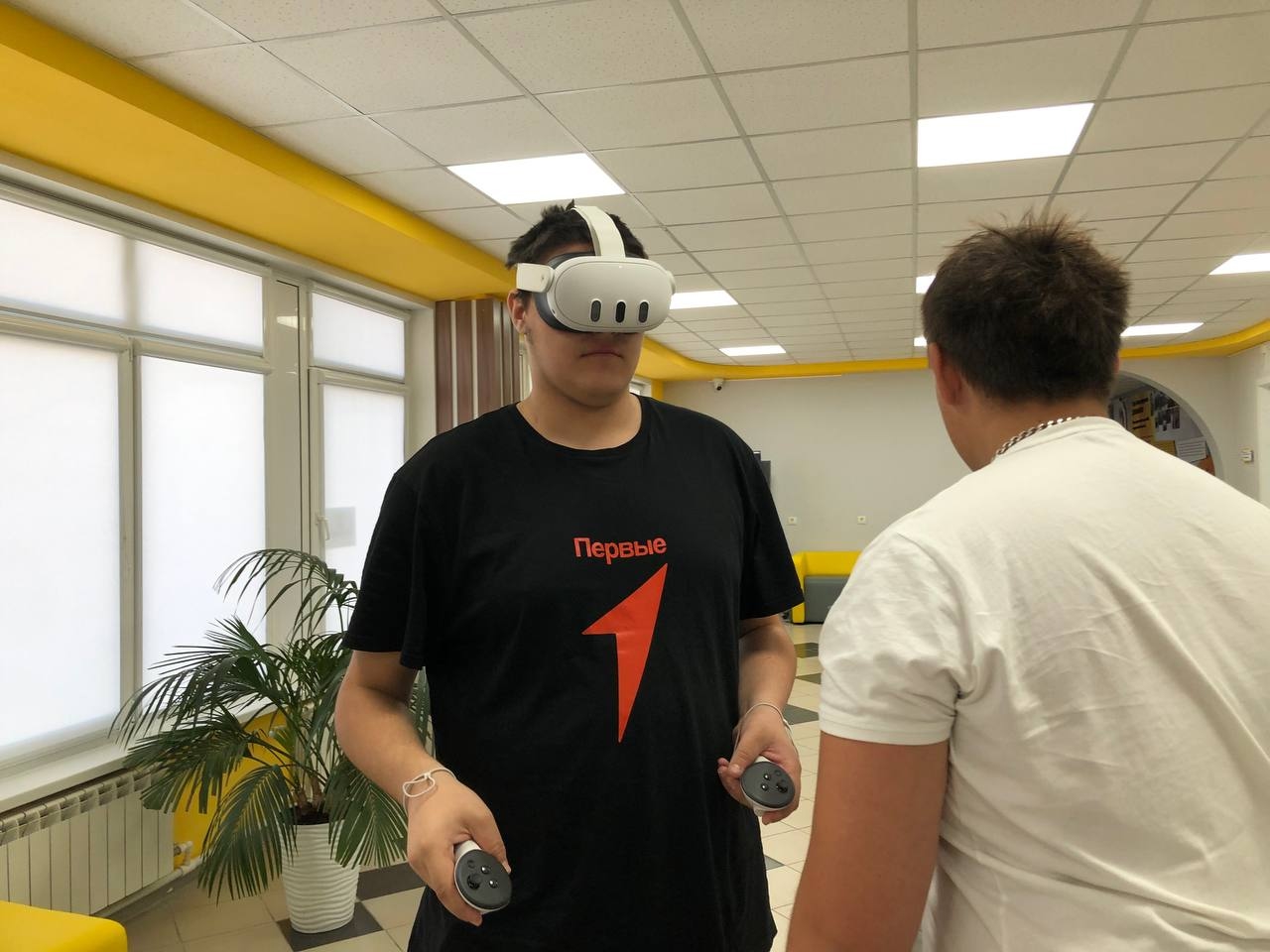 видно молодого человека, который носит VR-гарнитуру. Он одет в черную футболку с ярким логотипом, на нем держит два контроллера. Вокруг него расположены желтые и белые предметы интерьера, а также видно растение в углу. Вместе с ним находится другой человек, который смотрит на него.