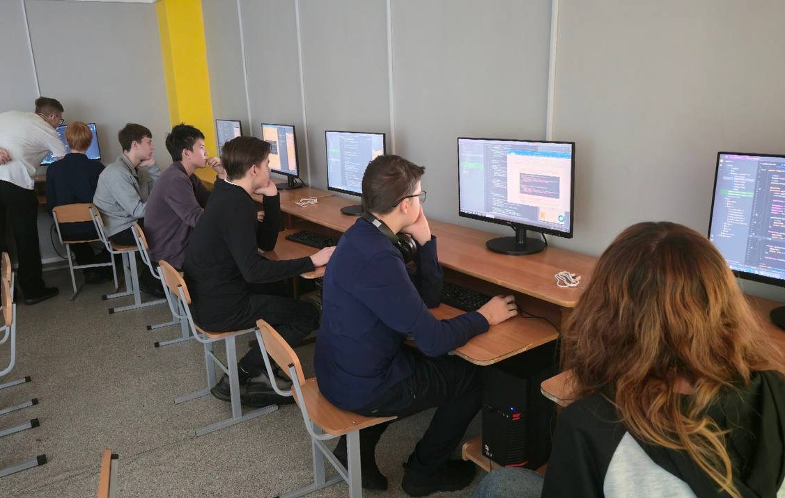 На изображении видно несколько человек, сидящих за столами в компьютерном классе. Все участники сосредоточены на экранах компьютеров, где открыты различные программы. В центре стола виден человек с наушниками, активно печатающий. Стены класса окрашены в серый цвет, а одна из них ярко-желтая. Освещение яркое, создающее рабочую атмосферу.