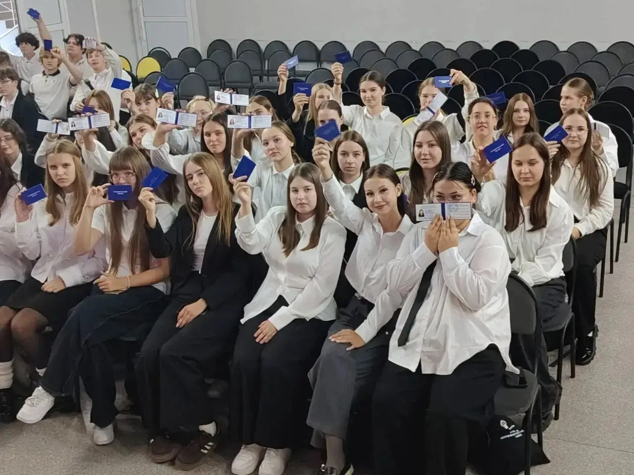 Групповое фото студентов, сидящих в несколько рядов в зале. Молодые люди и девушки одеты в парадную одежду: белые рубашки, блузки, темные юбки и брюки. Они смотрят в камеру и держат в поднятых руках синие студенческие билеты. На переднем плане девушка с темными волосами держит билет обеими руками near лица, рядом с ней улыбающаяся студентка подняла билет вверх. На заднем плане видны пустые ряды черных стульев. Атмосфера радости и торжественности.