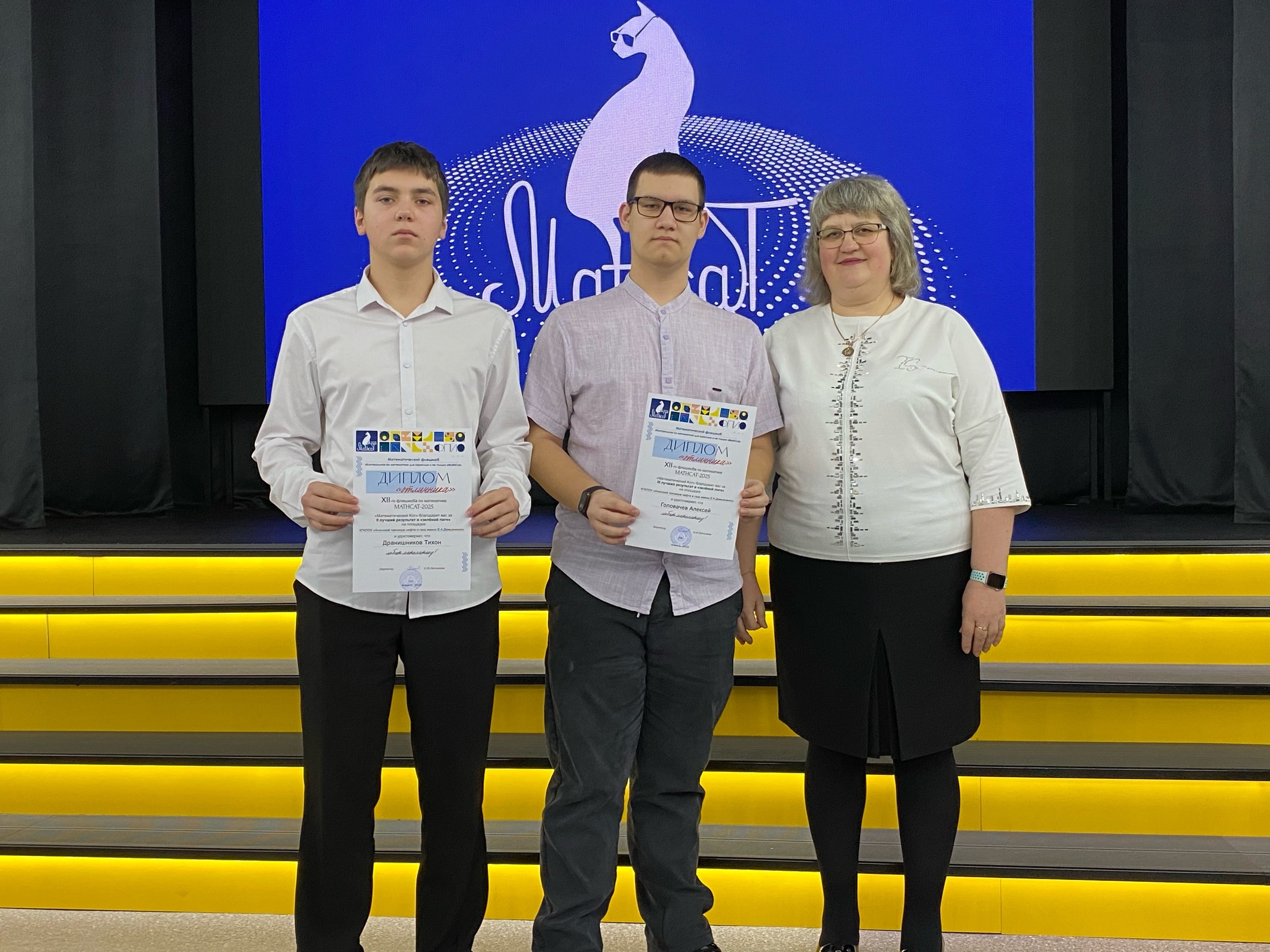 На сцене стоят три человека. Слева - молодой человек в белой рубашке с длинными рукавами, который держит диплом. В центре - ещё один молодой человек, также в белой рубашке, с короткими темными волосами и очками, держащий диплом. Справа - женщина в светлой блузке с длинными рукавами и светлыми волосами, она тоже держит диплом. На заднем плане видно большое синее оформление с логотипом мероприятия.