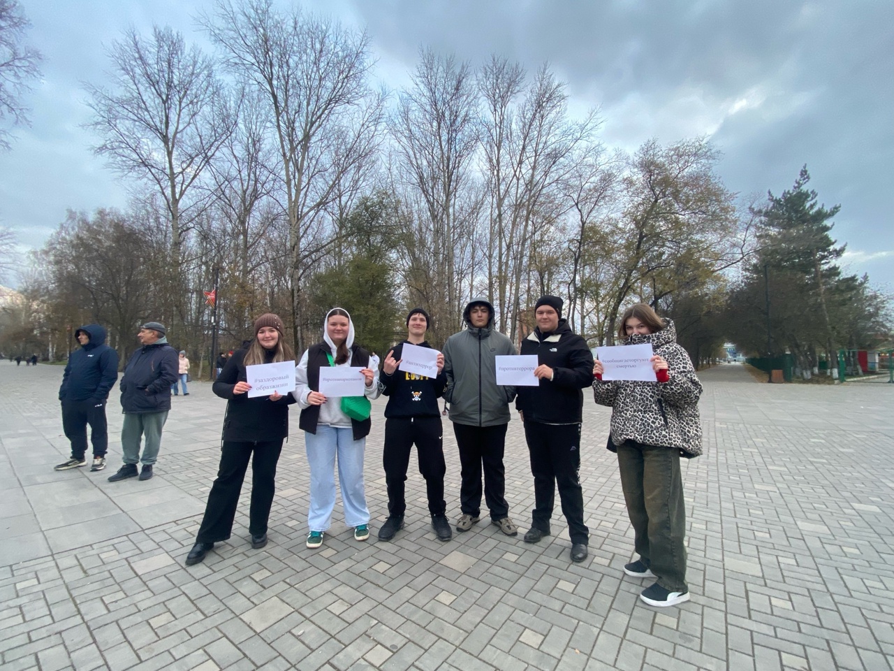 группа людей стоит на асфальтированной площадке. Они держат в руках белые листы бумаги. На заднем плане видны деревья, а небо затянуто облаками. Люди одеты в темные и тёплые одежды, также среди них есть один человек в ярком пиджаке.