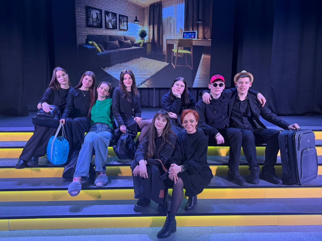 группа из девяти молодых людей сидит на ступенях, ведущих к сцене. Они одеты в чёрную одежду, несколько человек держат сумки. В фоне виден экран с изображением комнаты, где есть диван, кофейный столик и окно с солнечным светом. Атмосфера дружелюбная и непринуждённая.