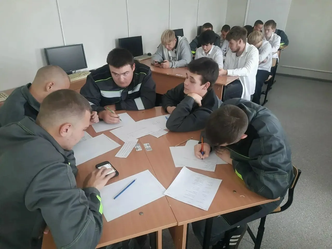Группа студентов в серых комбинезонах с желтыми полосами сидит за столом в аудитории. Перед ними разложены листы бумаги, ручки и смартфоны. Один студент пишет, другой облокотился на стол и смотрит в лист. На заднем плане другие студенты в белых рубашках, некоторые смотрят в телефоны. На столах стоят мониторы компьютеров. Освещение искусственное.