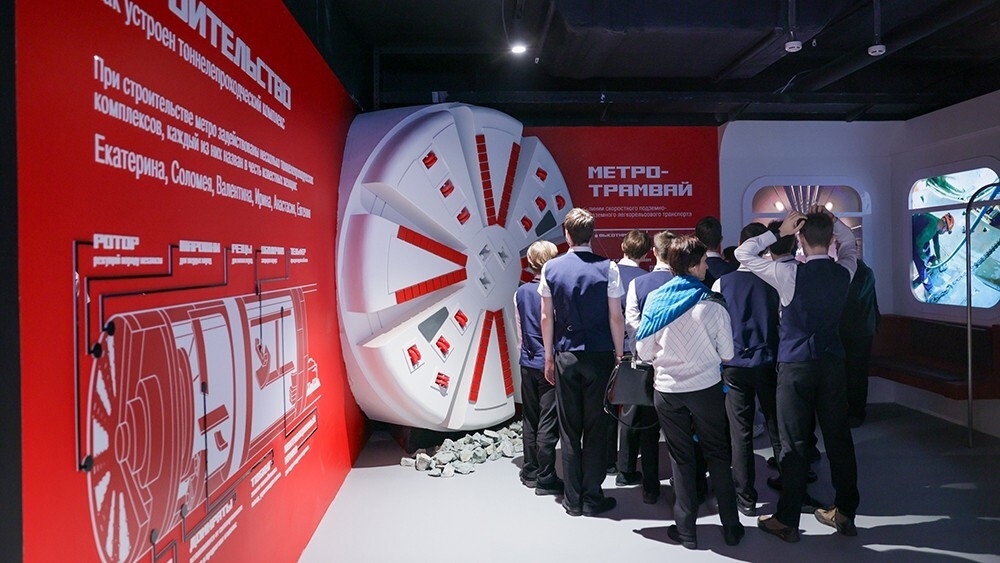 На фото изображена группа людей, стоящих перед огромным круглым объектом, напоминающим конструкцию для строительства метрополитена. Объект установлен на стенде с ярким красным текстом, который информирует о различных аспектах процесса создания метро. На стенде представлены схемы. Люди, одетые в формальные костюмы, внимательно слушают экскурсовода, который объясняет детали, стоя у объекта.