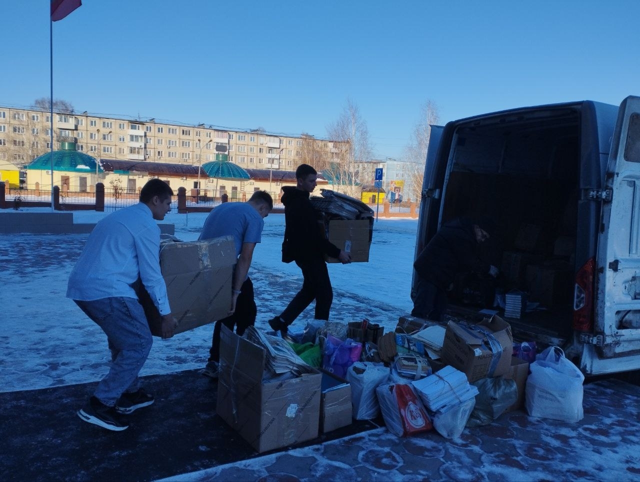 На изображении группа молодых людей, которые занимаются погрузкой коробок в белый фургон. На переднем плане видно, как двое человек переносят большие коробки, а один человек находится внутри машины. Вокруг их окружает снежный пейзаж, здания на заднем плане указывают на городской ландшафт. Всё это происходит в ясный, солнечный день, на фоне голубого неба. На земле можно увидеть разбросанные пакеты и вещи, которые также пытаются загрузить в машину.