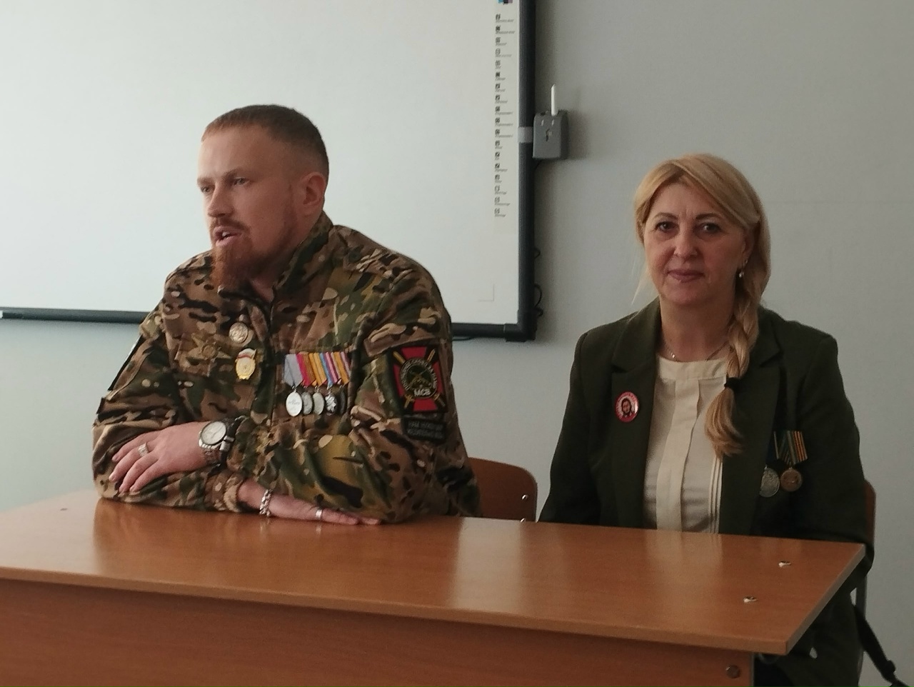 На этом фото мужчина в камуфляжной форме с множеством медалей сидит за столом, смотря в камеру. Рядом с ним сидит женщина, одетая в темное платье и костюм. Оба визуально сосредоточены и выглядят уверенными. В классе на стене виден экран, а также настенные панно.