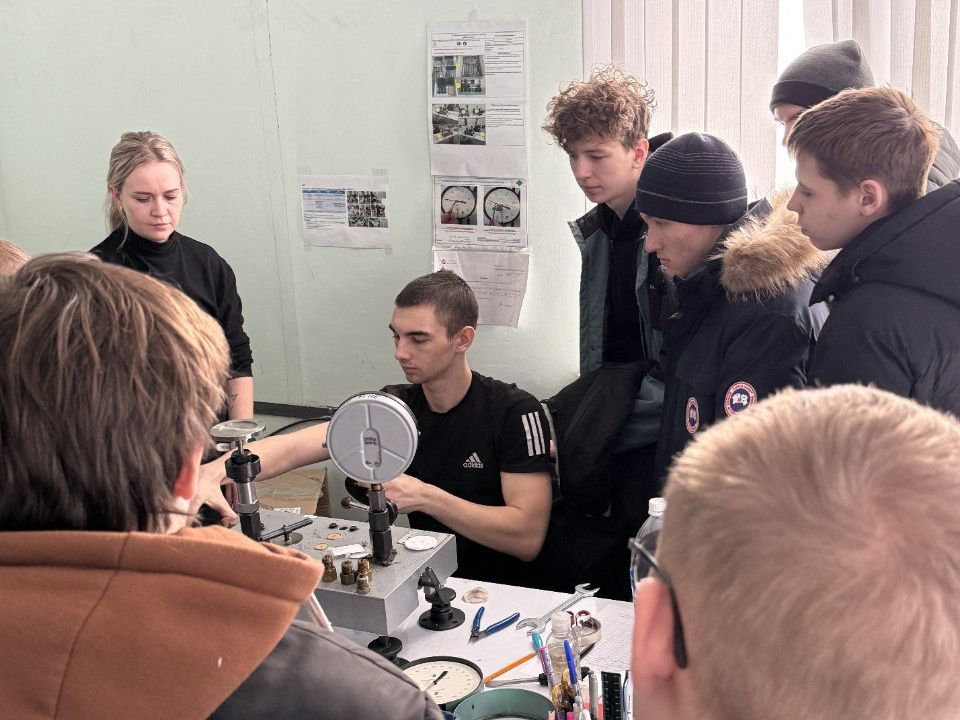 группа молодых людей, внимательно наблюдающих за процессом работы. В центре сидит парень, занимающийся техникой, вокруг него несколько человек, включая девушку, которая указывает на прибор. Все выглядят сосредоточенными. Стены помещения светло-зеленые, а на них висят учебные материалы и графики.