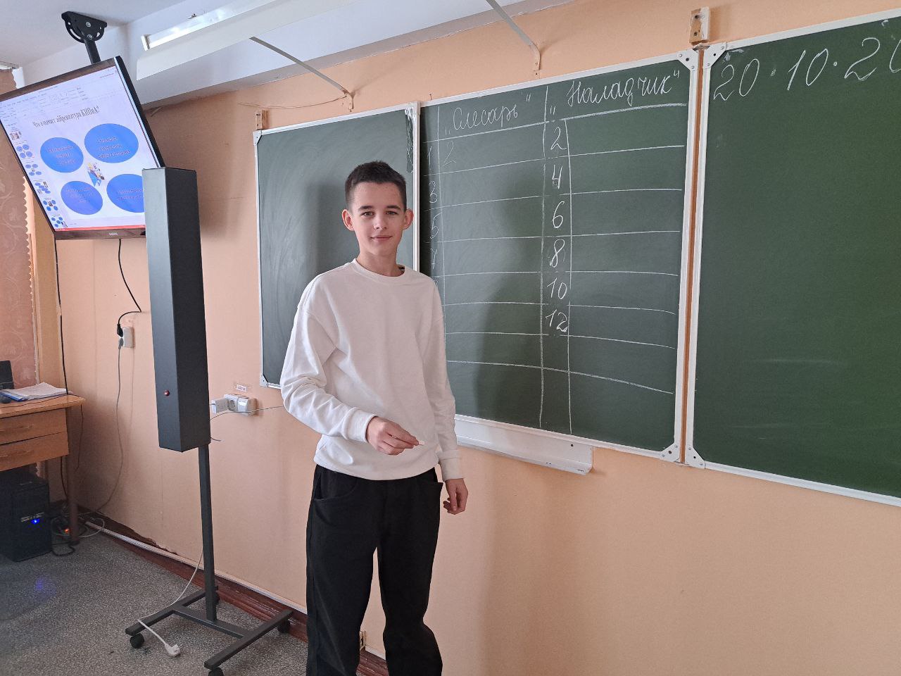 изображен мальчик, стоящий у доски в классе. Он одет в белую футболку и черные штаны. На заднем плане видна зеленая доска для письма и экран, на котором отображаются графические элементы. Мальчик, вероятно, готовится объяснять что-то классу.