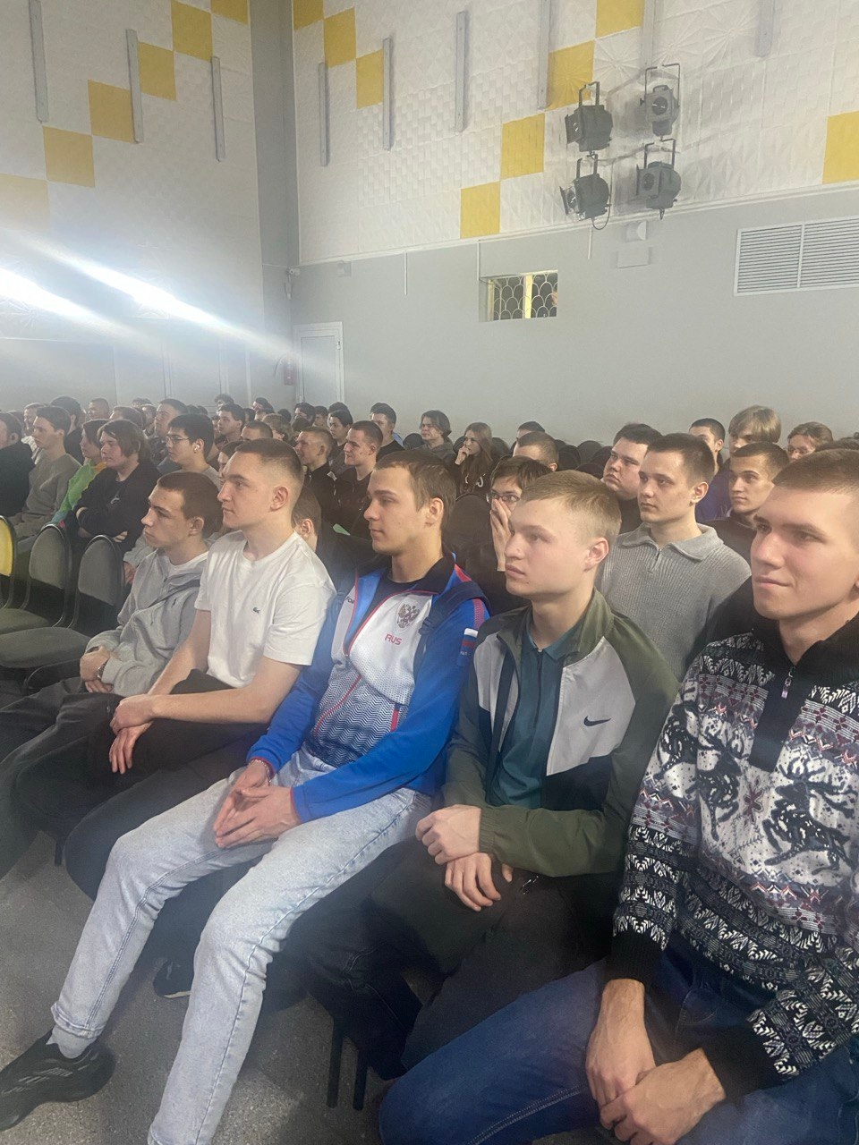 На картинке изображена большая группа людей, сидящих в зале. Все они смотрят в одной направлению, скорее всего, на сцену или экран. В центре внимания — молодые мужчины, большинство в светлой одежде. Мужчины разных возрастов, но преимущественно молодые, сидят в рядах на черных стульях. Стены зала окрашены в светлые тона, есть элементы декора в виде желтых квадратов. Освещение достаточно яркое, создающее атмосферу учебного или делового мероприятия.