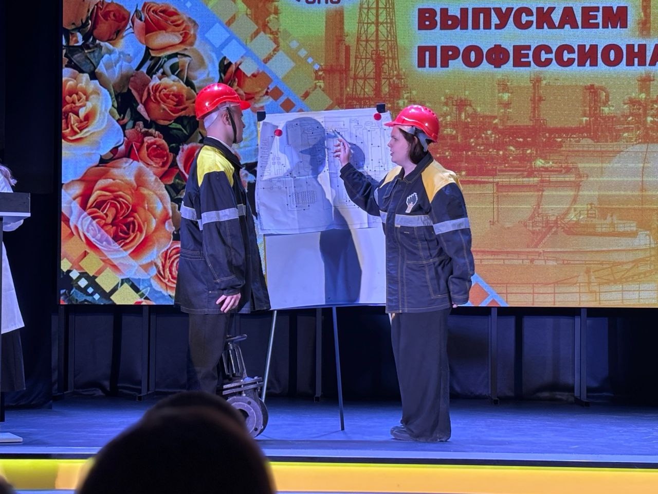 два человека, одетых в защитную форму и каски. Они стоят перед большим листом бумаги с чертежом. Один из них указывает на чертеж, объясняя детали. На заднем плане — яркий баннер с надписью "Выпускаем профессионалов", обрамленный цветами, что также разделяет сцену на цветные зоны.