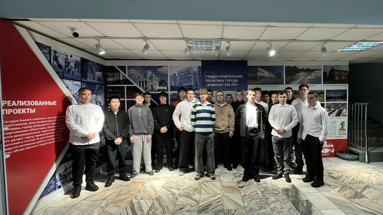 представлена группа из молодых людей, стоящих на фоне стенда с информацией о реализованных проектах города. Все они одеты в белые и серые свитера, некоторые в спортивной одежде. В центре стоит молодой человек в полосатой рубашке, который выглядит уверенно. Окружающие стены содержат фотографии зданий и планы развития городских проектов, создавая интересный контекст для группы.